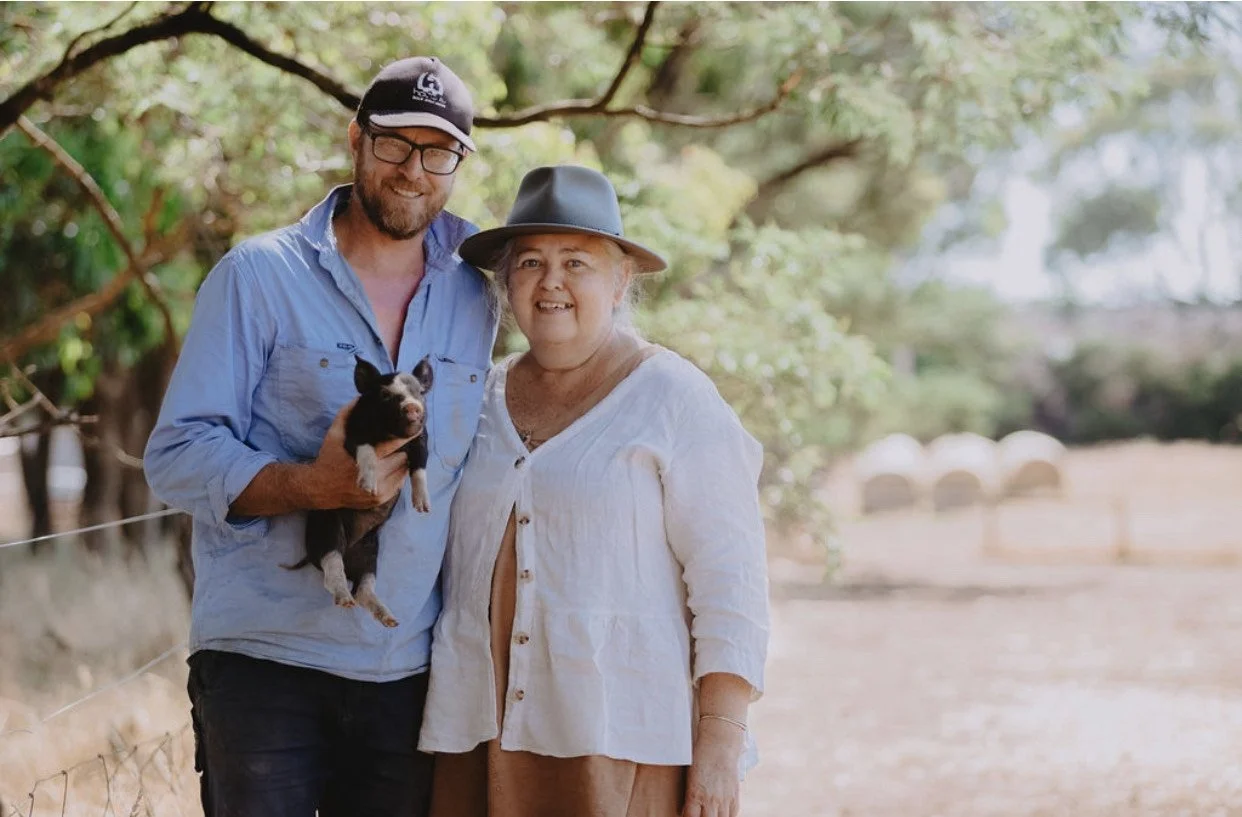 A man and woman standing outdoors, the man holding a small black and white piglet, in a rural setting with trees and a fence in the background.