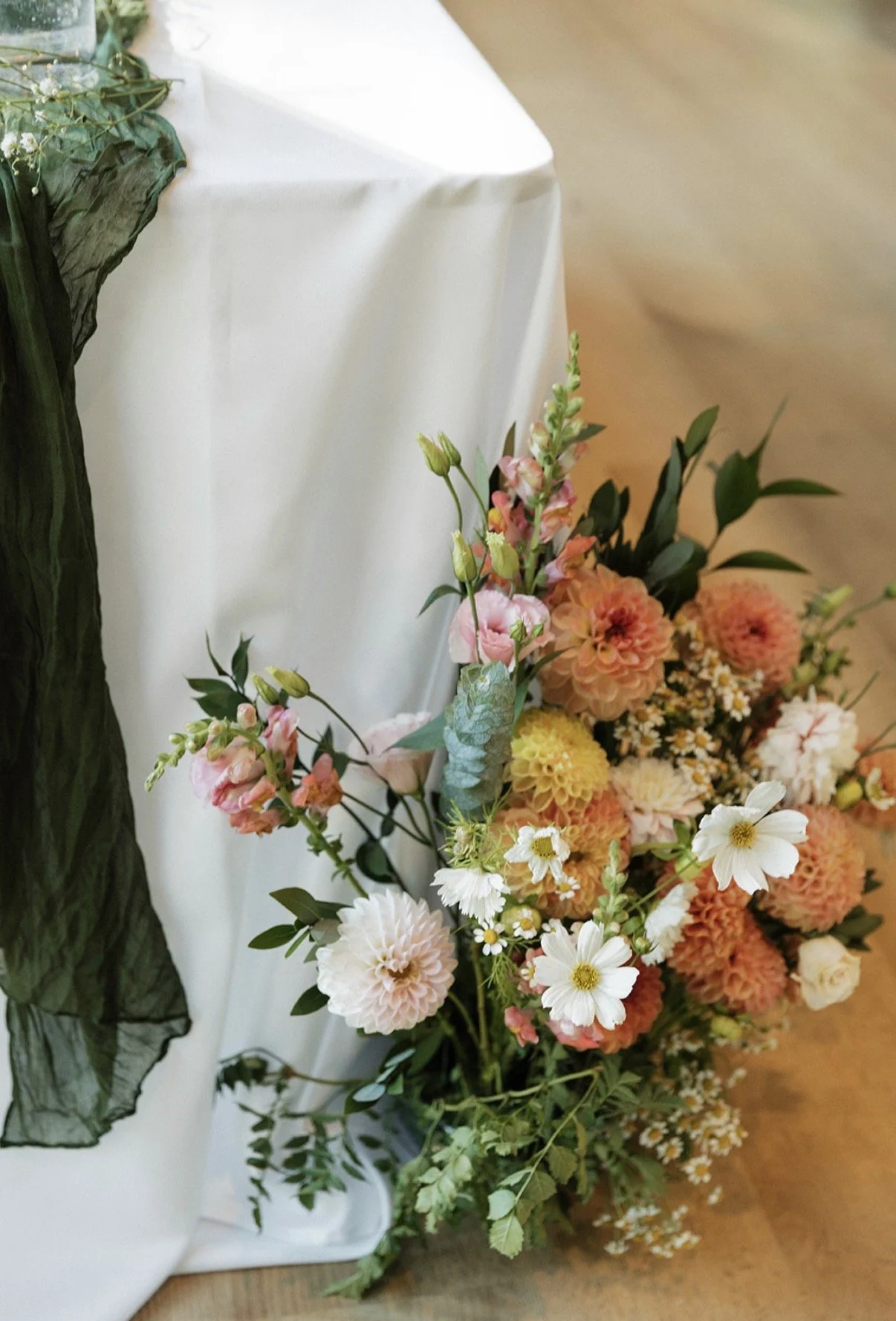 A young girl in a floral dress sitting outdoors, blowing on a dandelion puff.