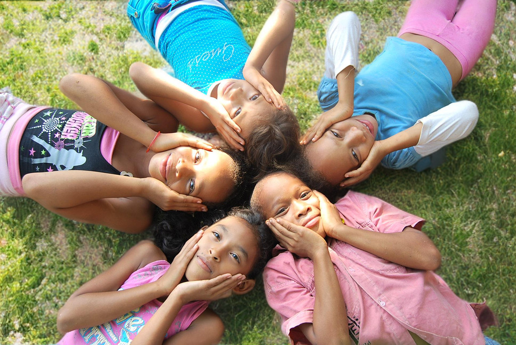Cinco niños recostados en el césped formando un círculo, mirando hacia arriba y sosteniendo sus rostros con las manos.