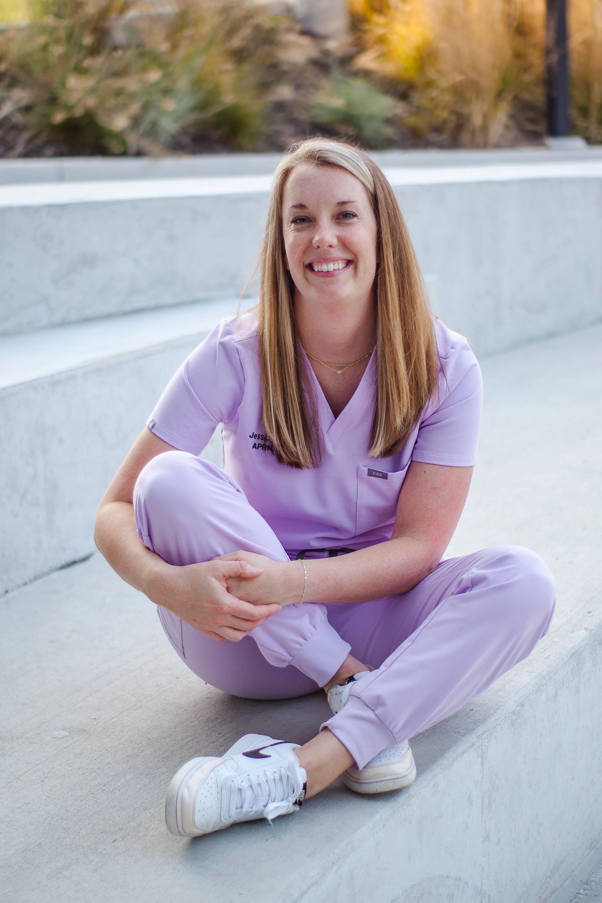 A smiling woman with long blonde hair sitting cross-legged on concrete stairs outdoors, wearing lavender medical scrubs and white Nike sneakers.