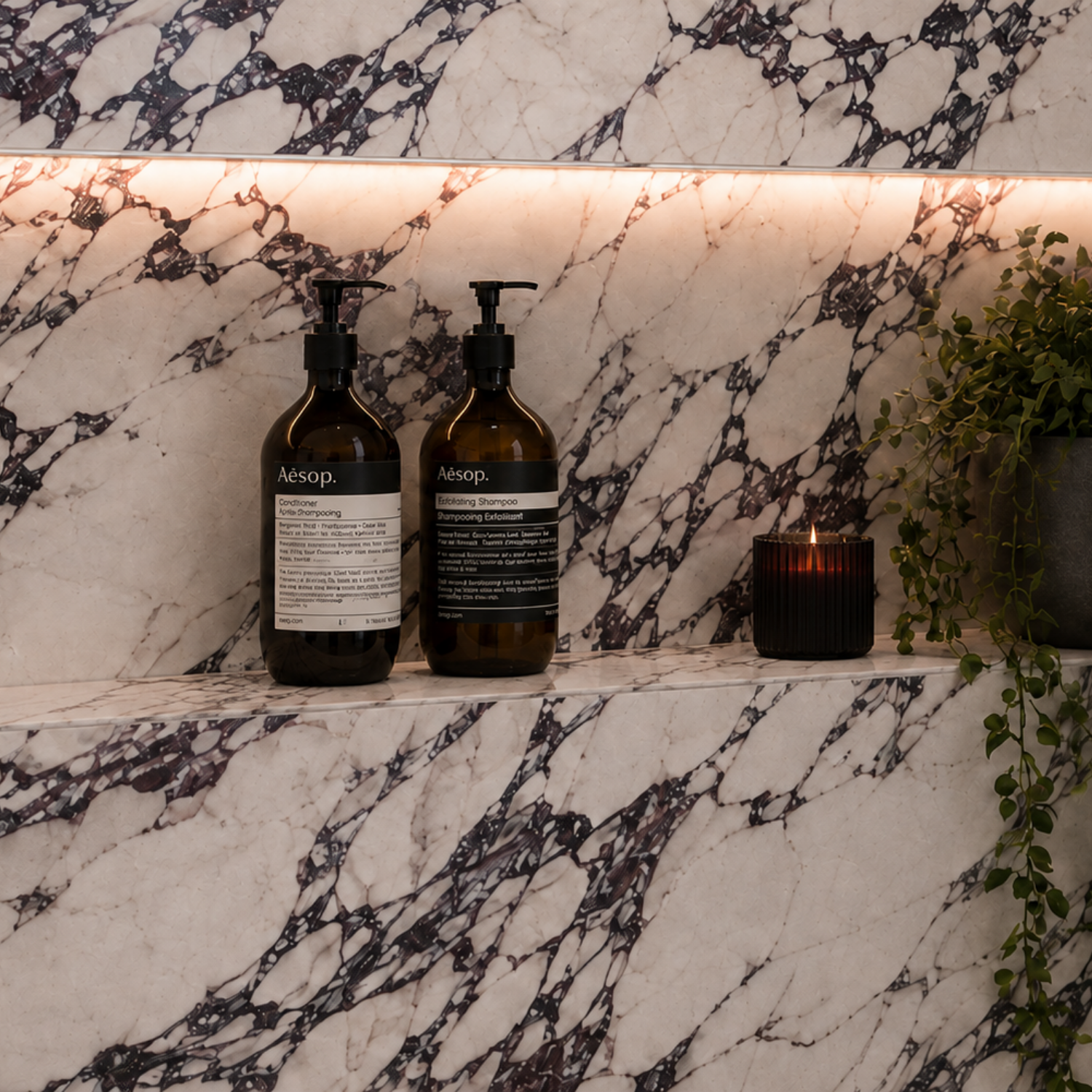 Close-up of Arabesco Viola marble-look porcelain bathroom wall tiles with dramatic dark veining, a softly illuminated recessed niche, amber bottles and greenery.