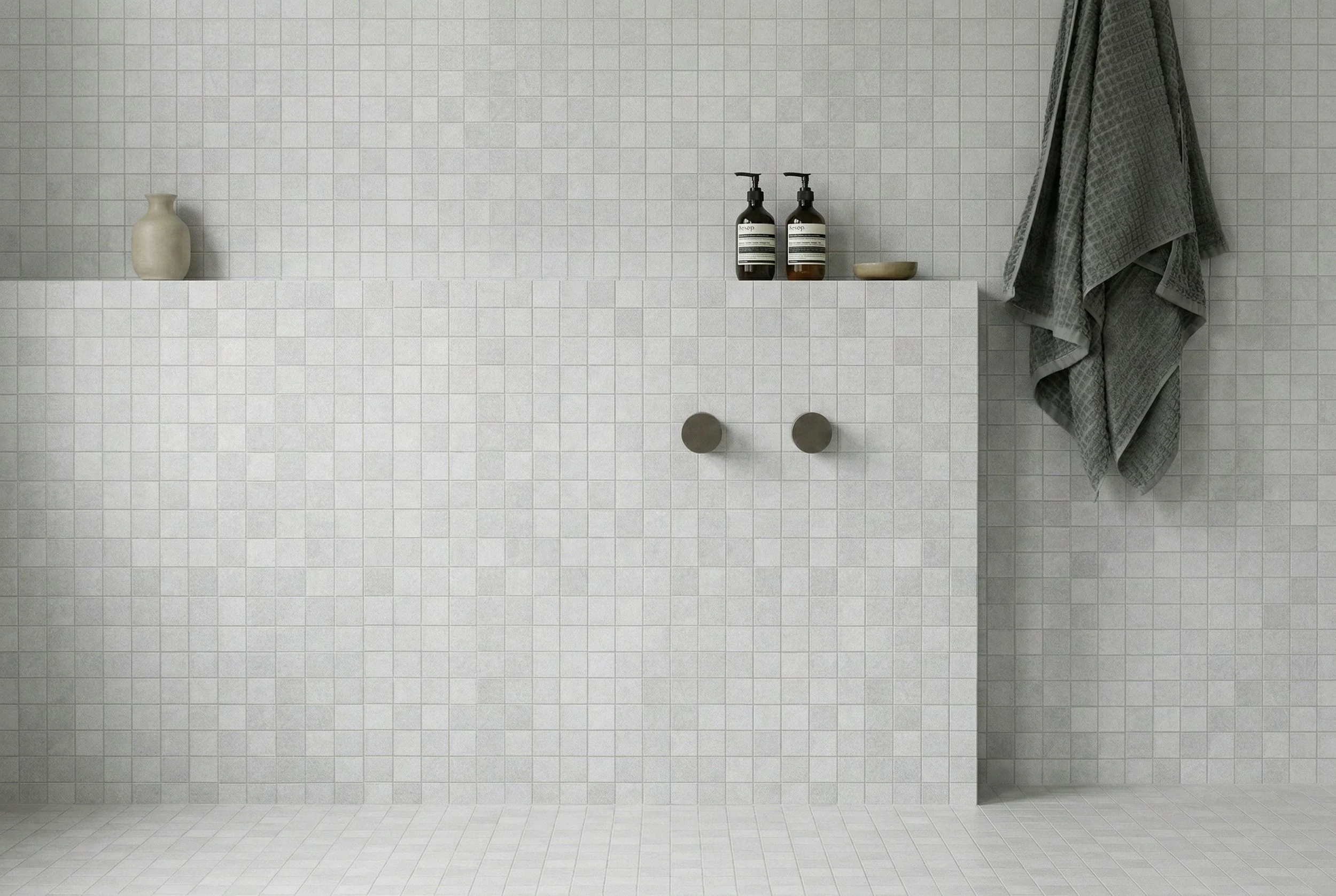 Bathroom featuring Limestone Tumbled Mist grey porcelain mosaic tiles across the walls and floor, with a recessed shelf, dark tapware, amber soap bottles and a hanging grey towel.
