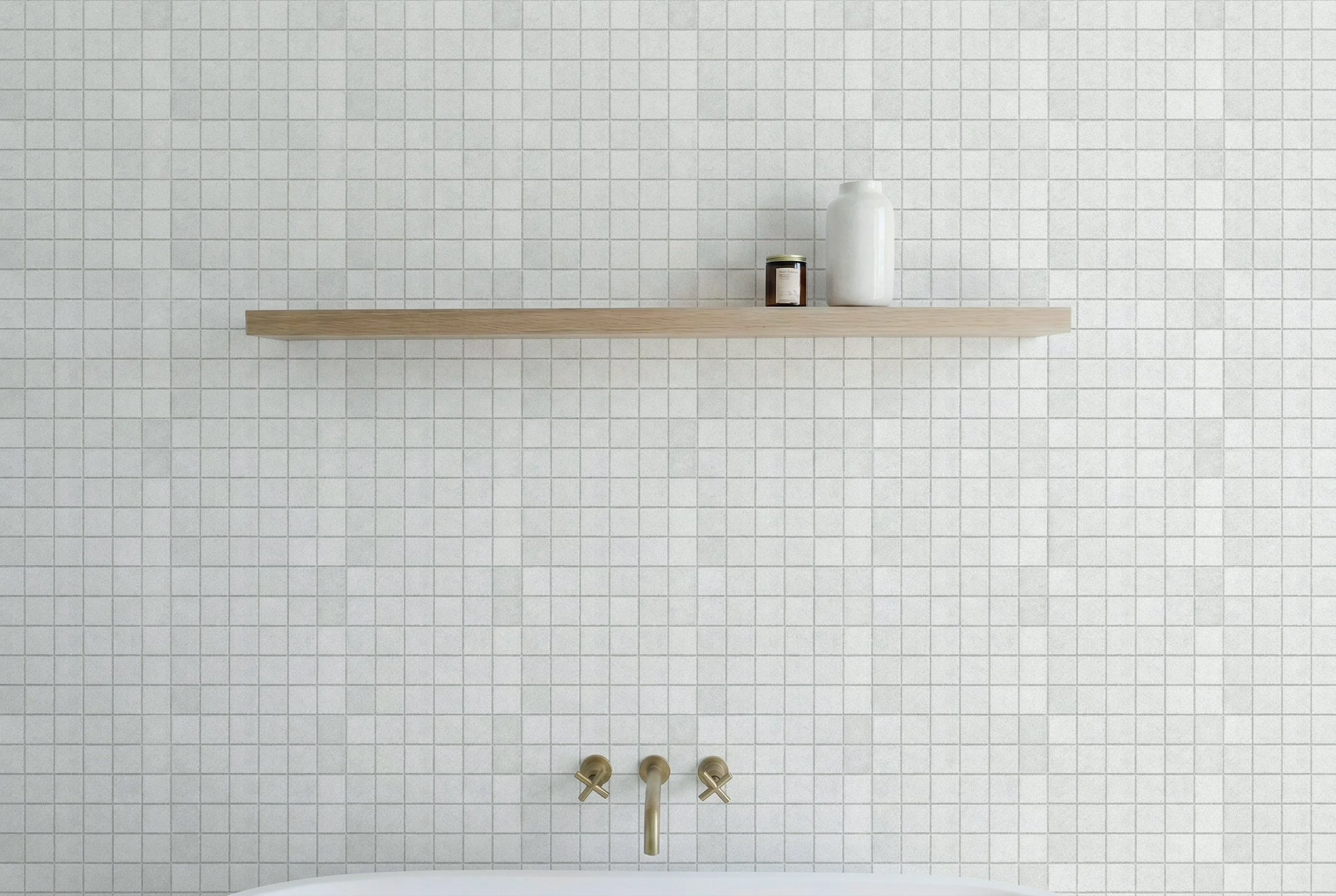 Bathroom wall featuring Limestone Tumbled Bianco off-white square porcelain mosaic tiles, a floating timber shelf, white accessories and brass tapware above a bathtub.