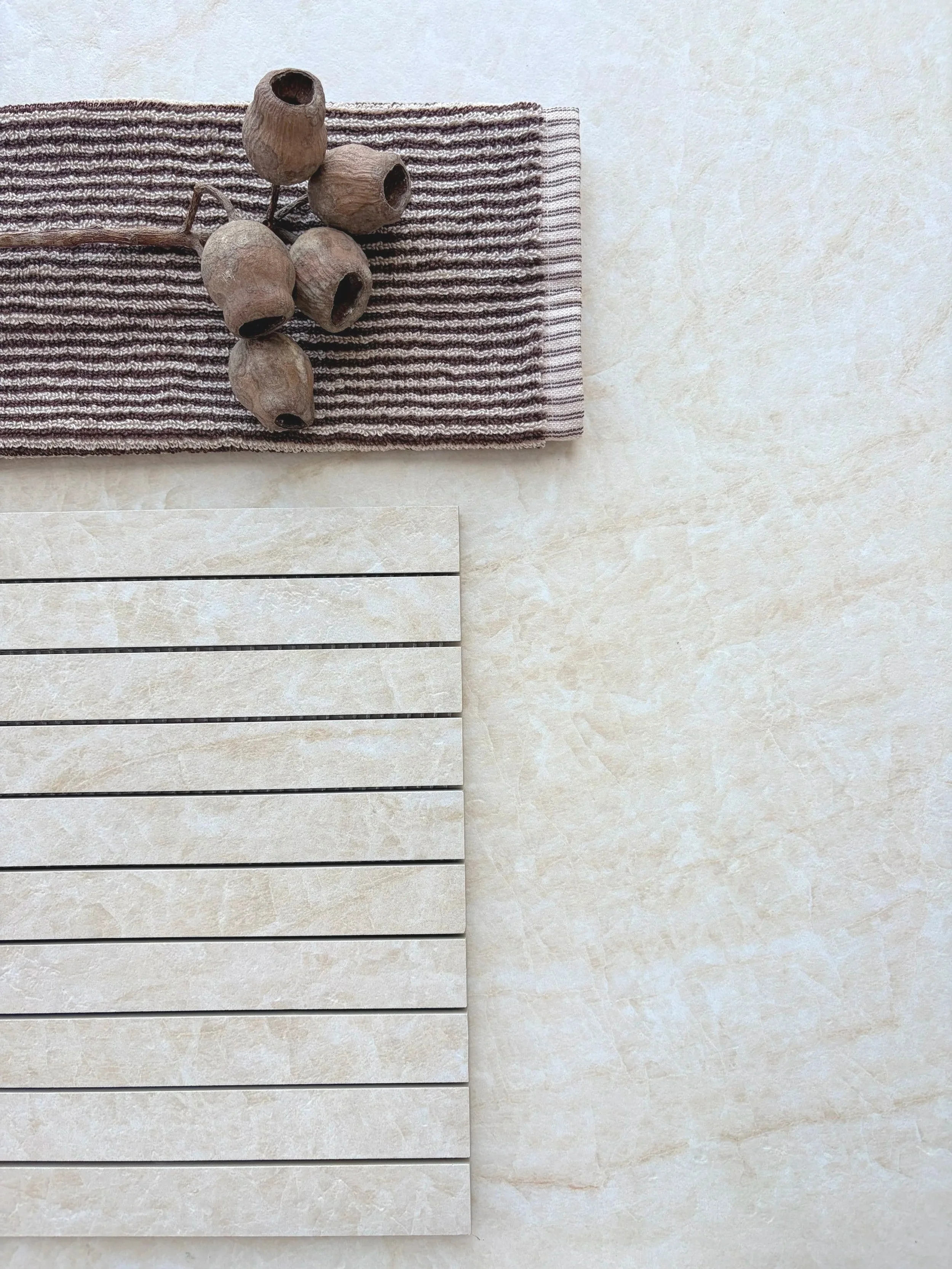 Taj Mahal Linea porcelain mosaic strips arranged horizontally beside a warm cream tile surface, striped fabric and dried seed pods.