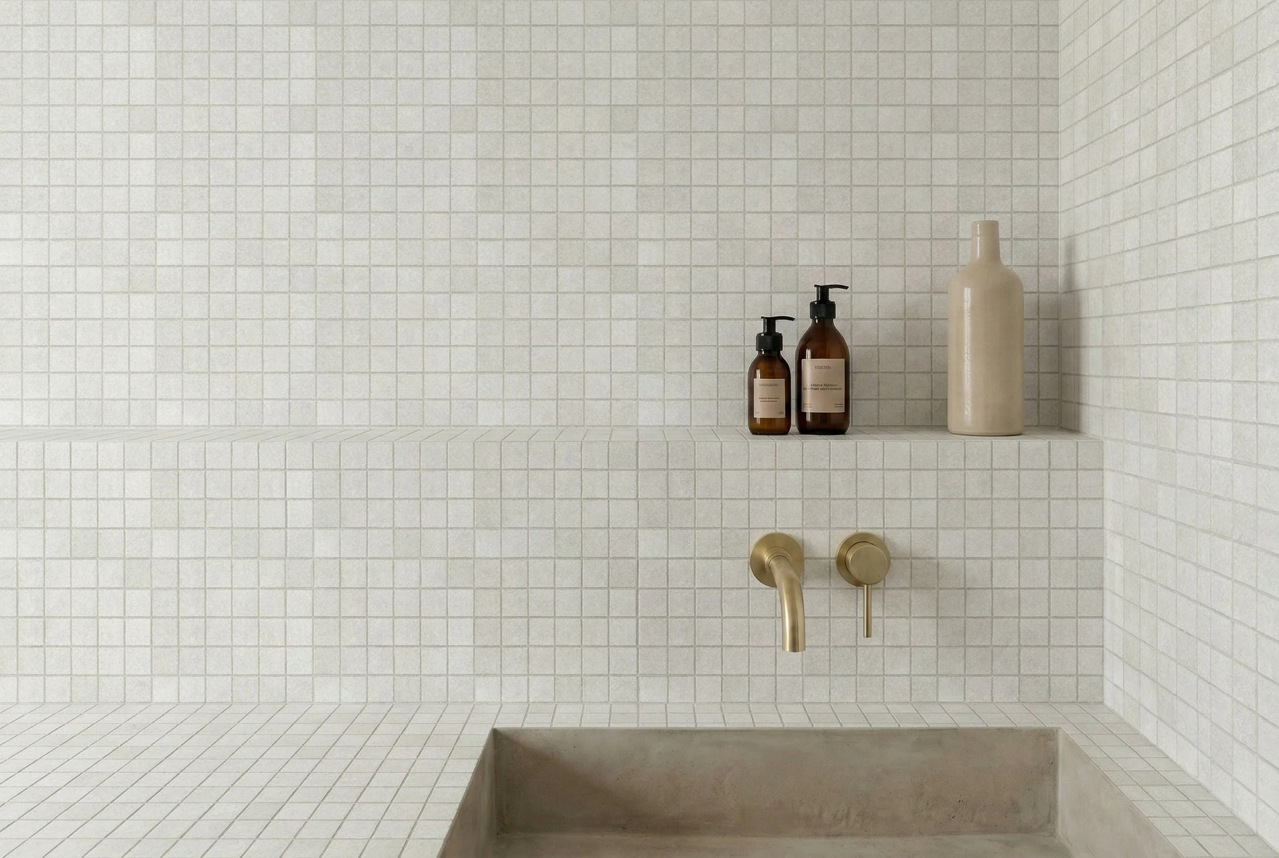 Bathroom featuring Limestone Tumbled Ivory square porcelain mosaic tiles across the walls and bath surround, with a recessed shelf, brass tapware and neutral accessories.
