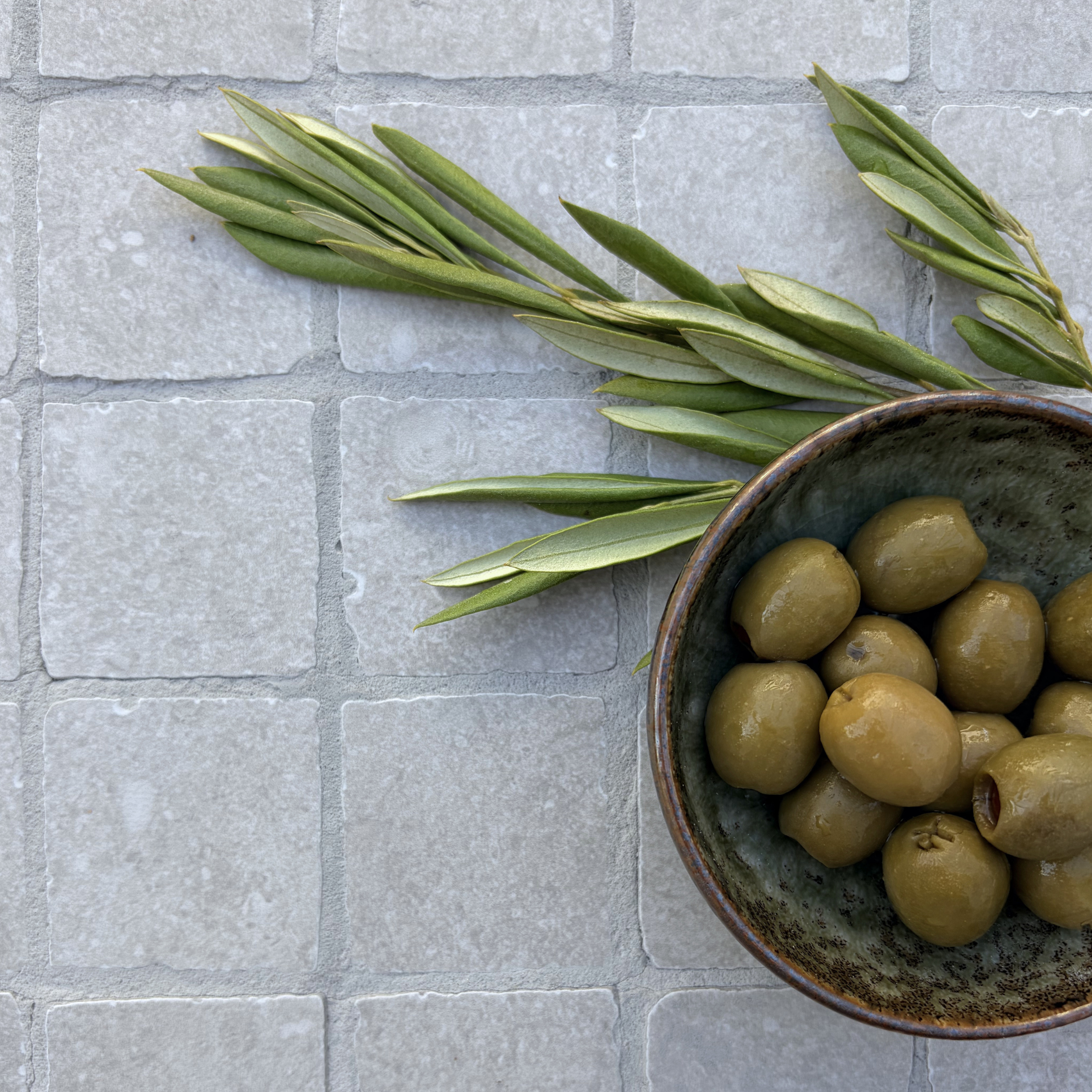 Limestone Tumbled Mist grey porcelain mosaic tiles with softened square edges, styled with a rustic ceramic bowl of green olives and fresh olive branches.