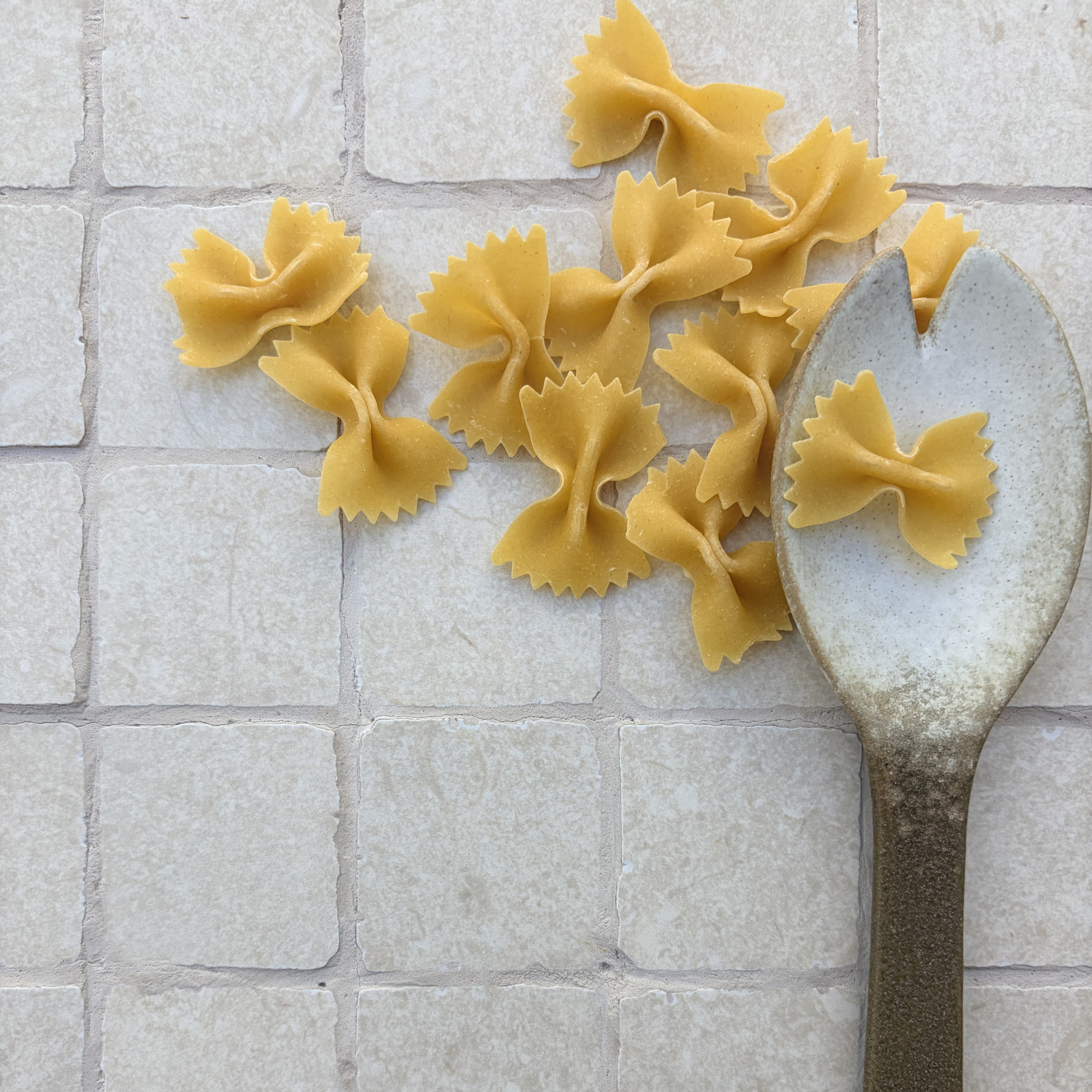 Limestone Tumbled Ivory porcelain mosaic tiles with soft ivory tones and irregular tumbled edges, styled with scattered bow-tie pasta and a rustic metal spoon.