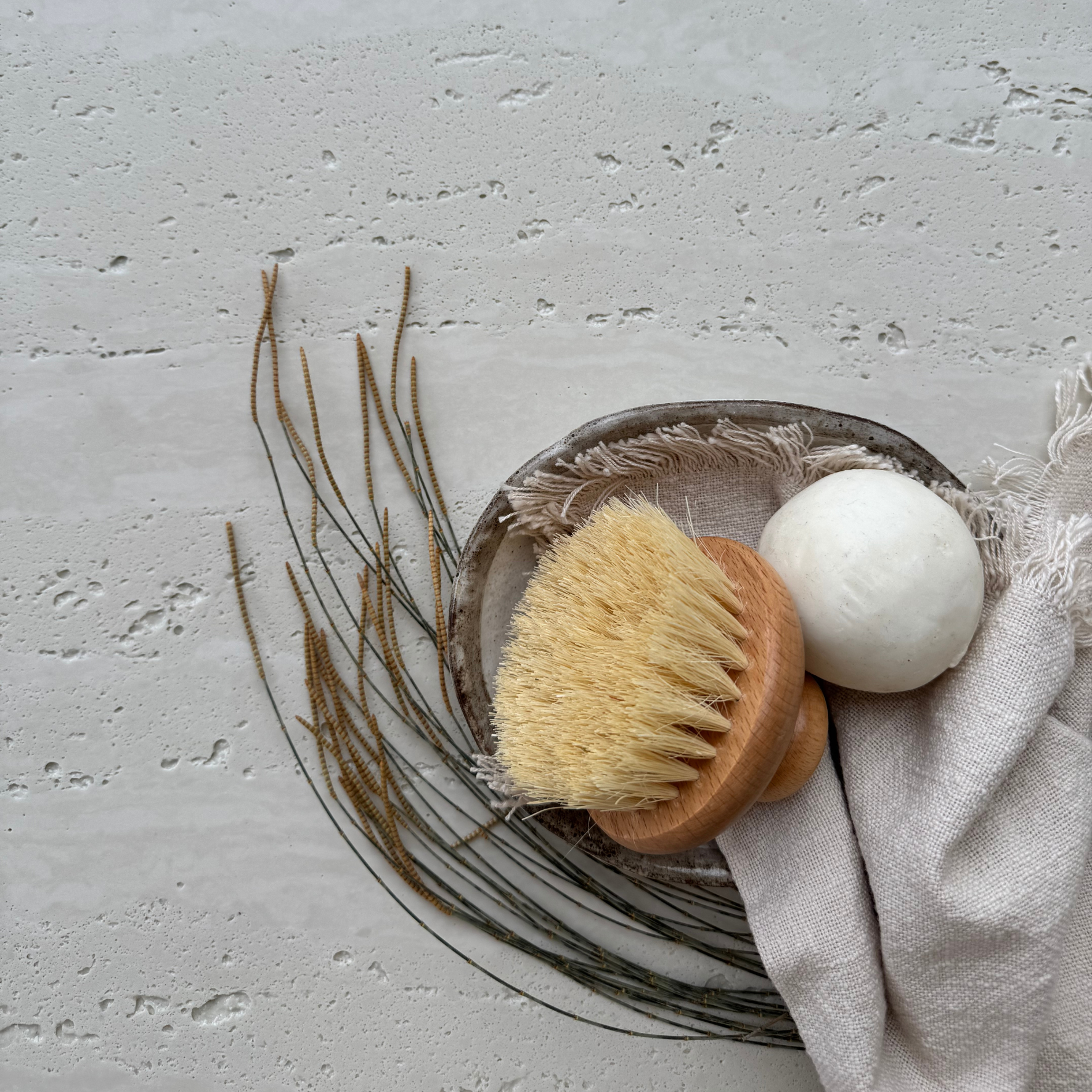 Travertine Classico Linen porcelain tile styled with a wooden body brush, white soap, dried twigs and neutral linen fabric against its textured stone-look surface.