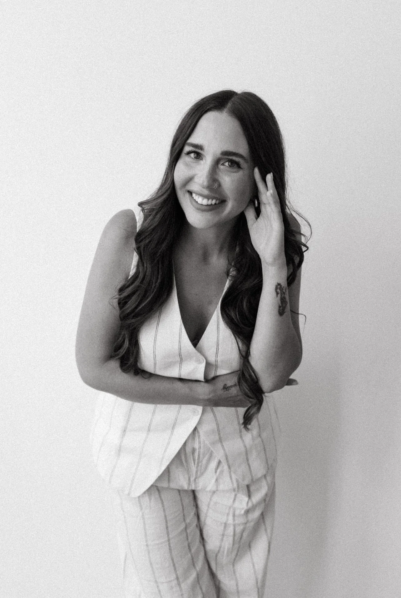 Black and white photo of a woman with long dark hair, smiling, wearing a sleeveless striped suit, standing against a plain light-colored wall.