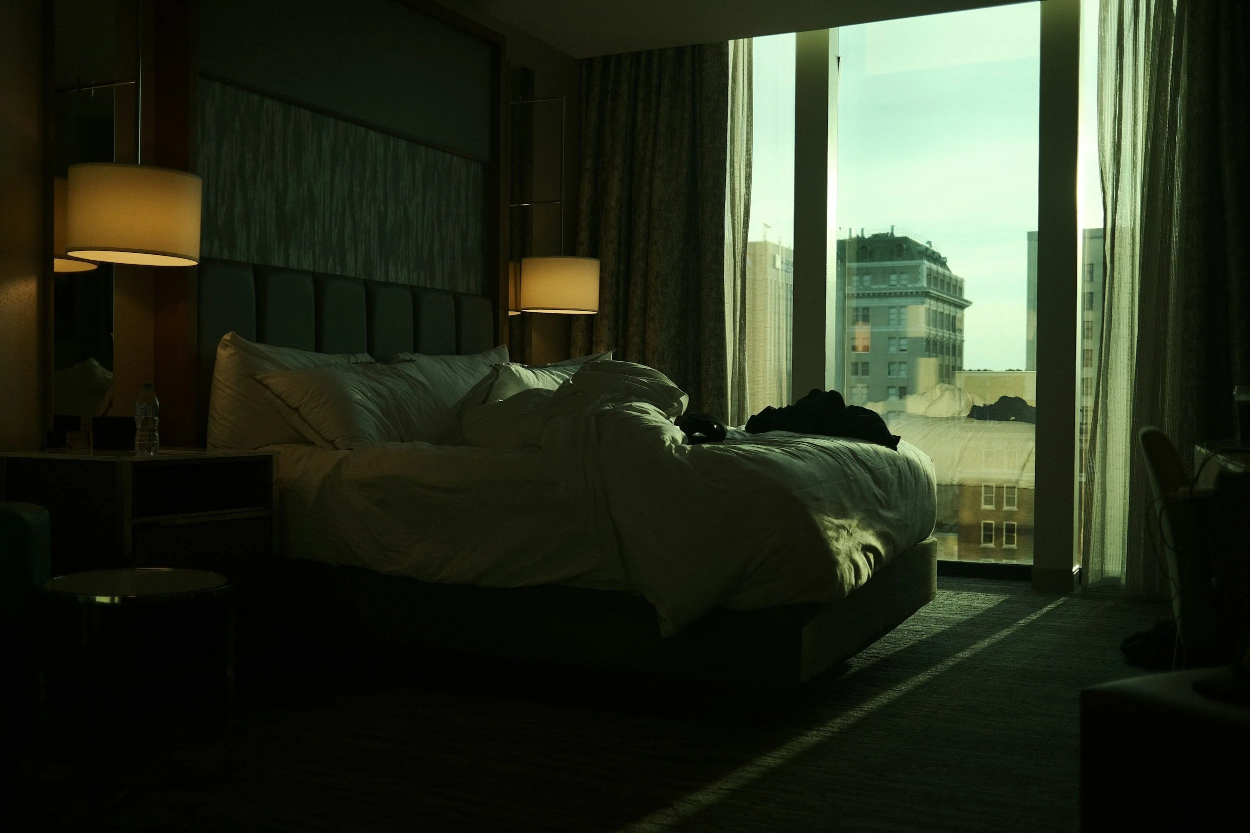 A dark hotel room with an unmade bed near large windows revealing city buildings outside. Bedding is rumpled and clothes are on the bed, with sunlight casting shadows inside.