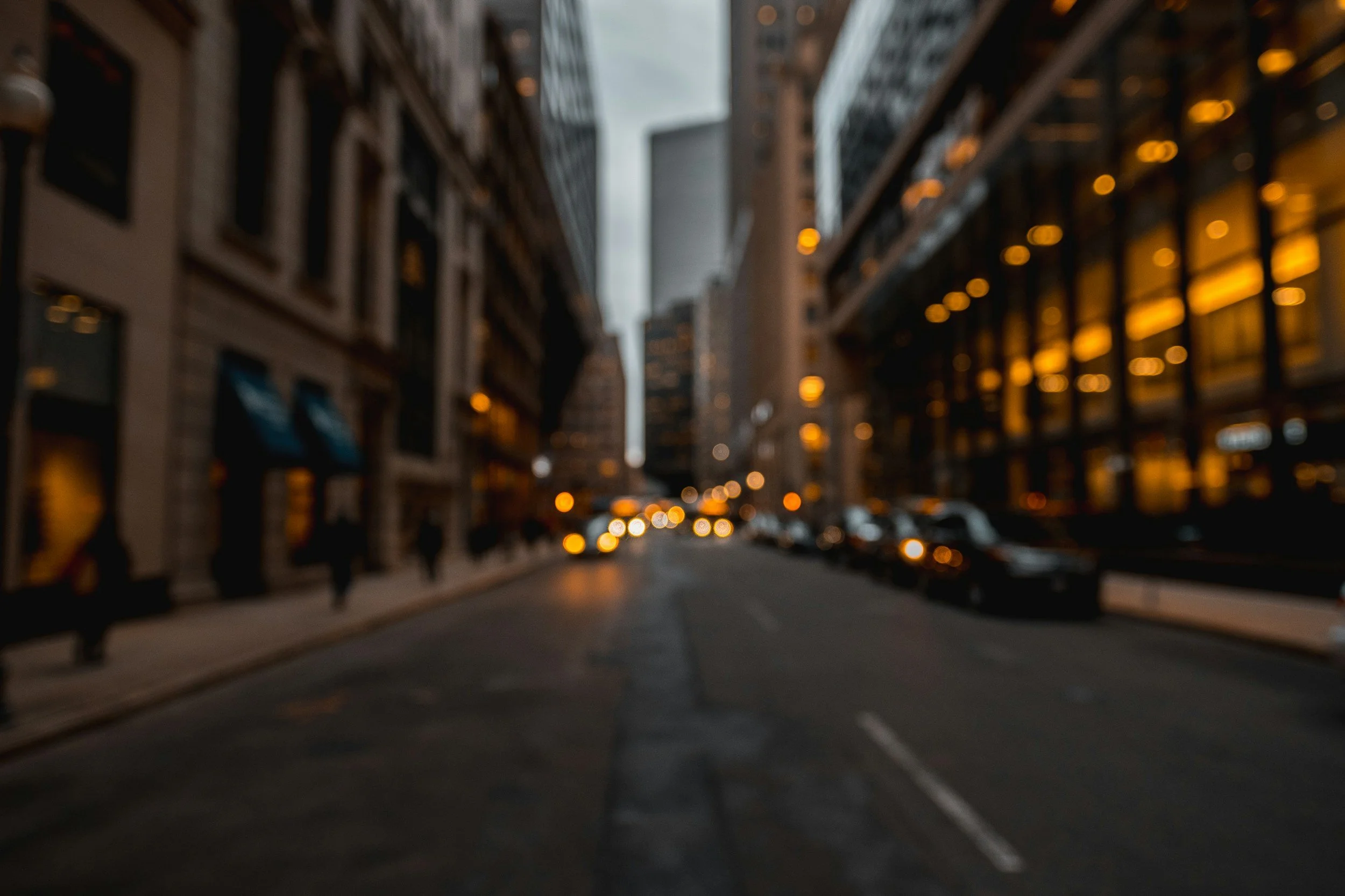 Blurred city street scene with tall buildings on both sides, cars parked along the curb, and pedestrians walking on the sidewalk. Evening lighting with some illuminated windows and streetlights.