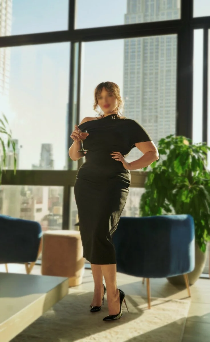 Zoey Belladonna in a black dress and high heels holding a glass of wine, standing in a modern high-rise apartment with large windows and skyline view of New York City, including tall buildings and some plants.