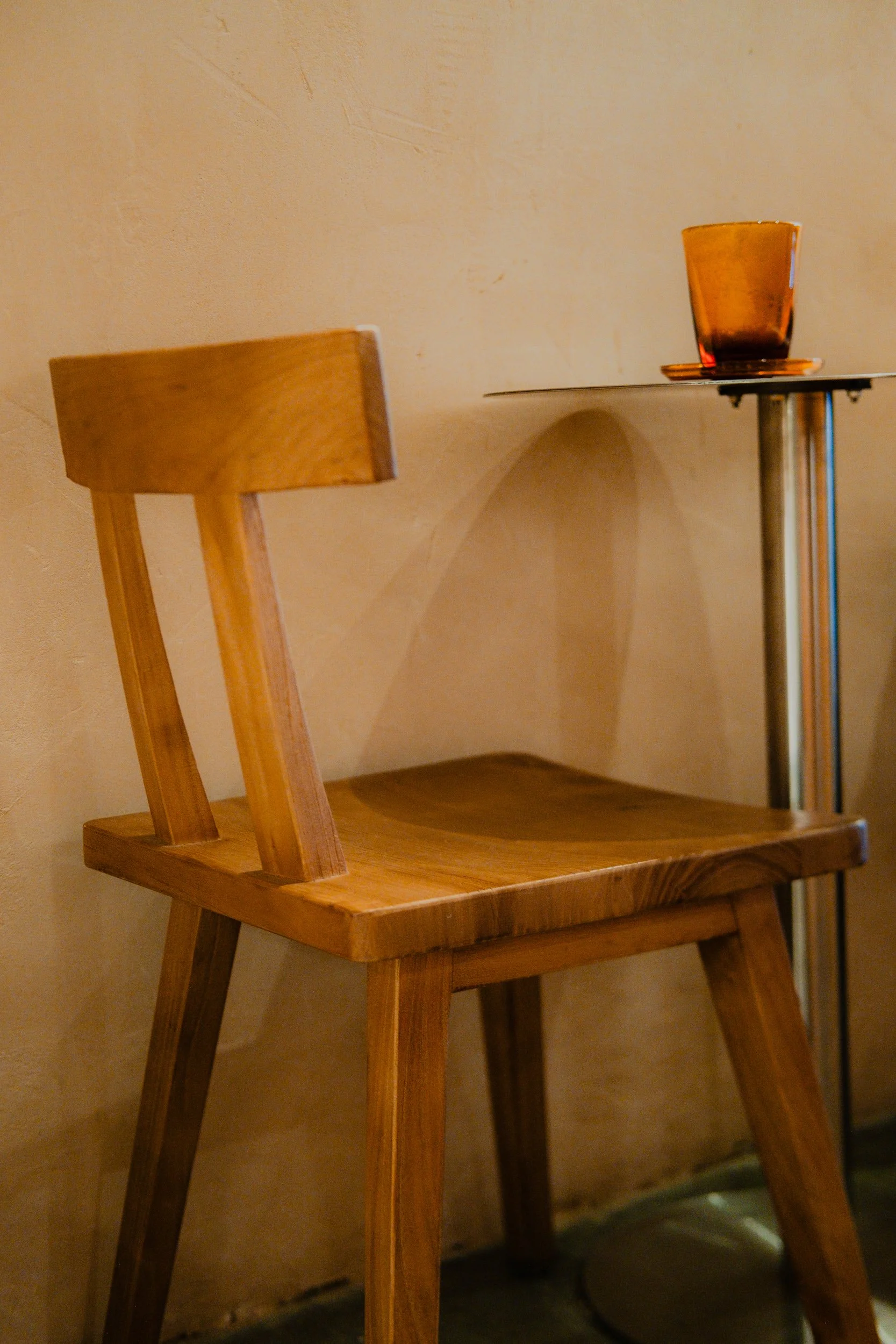 A wooden chair with a curved backrest next to a small table with a glass on it. The glass is amber-colored, and the scene appears to be indoors.