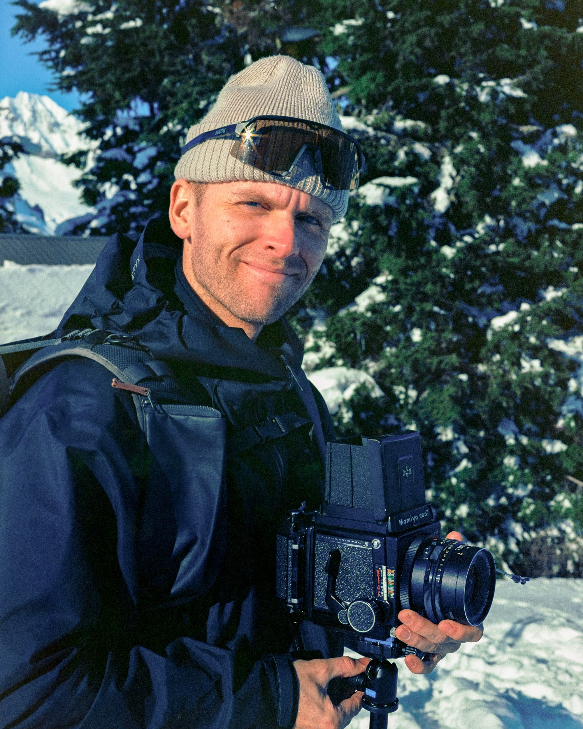 Photographer holding a camera on a tripod in a snowy landscape, wearing a knit hat and sunglasses, with evergreen trees and mountains in the background.
