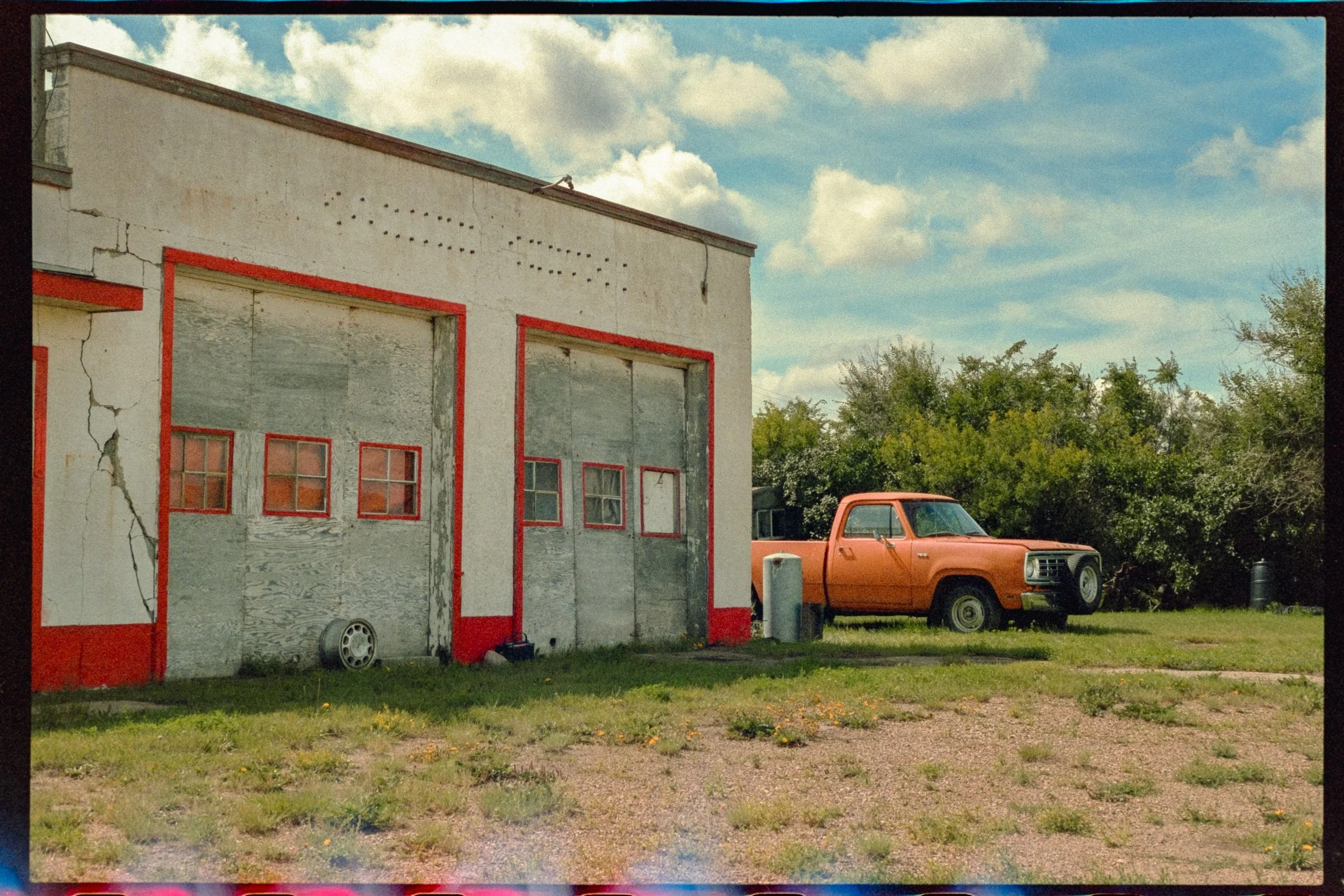 An old, worn-out garage building with cracked white walls and red trim. There are small windows on a large, boarded-up garage door. A rust-colored pickup truck is parked on the grass nearby, with green bushes and a partly cloudy sky in the background