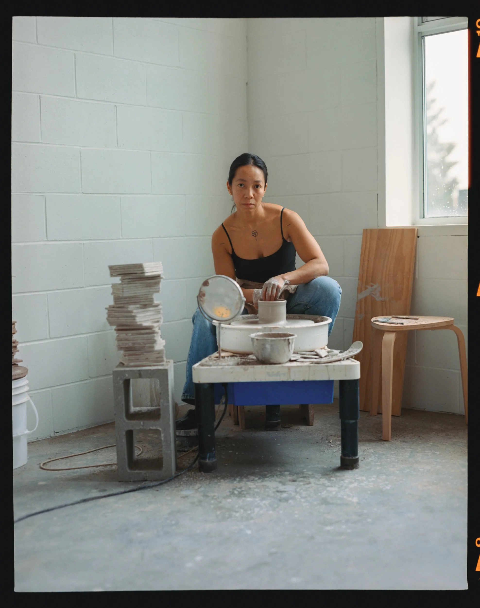 A woman working on pottery at a pottery wheel, surrounded by stacks of clay pieces and pottery tools in a studio with natural light from a window.
