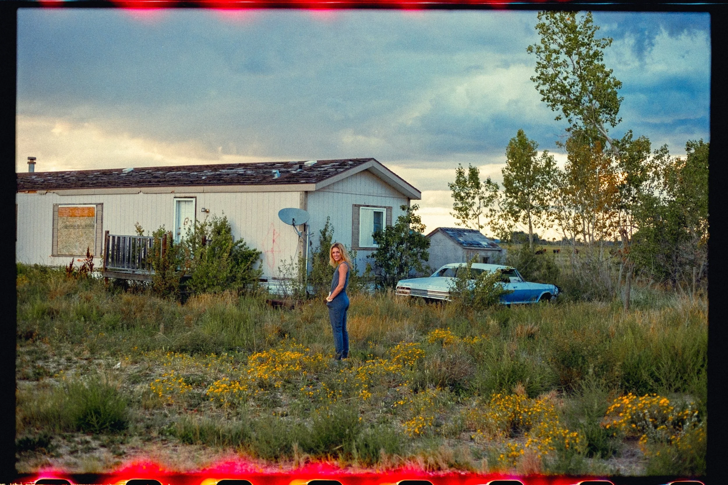 A woman in a tank top and jeans standing in a grassy, overgrown yard with yellow flowers, near an older beige house with a satellite dish and a small wooden deck. A classic blue and white car is parked near trees and bushes, and the sky is partly clo