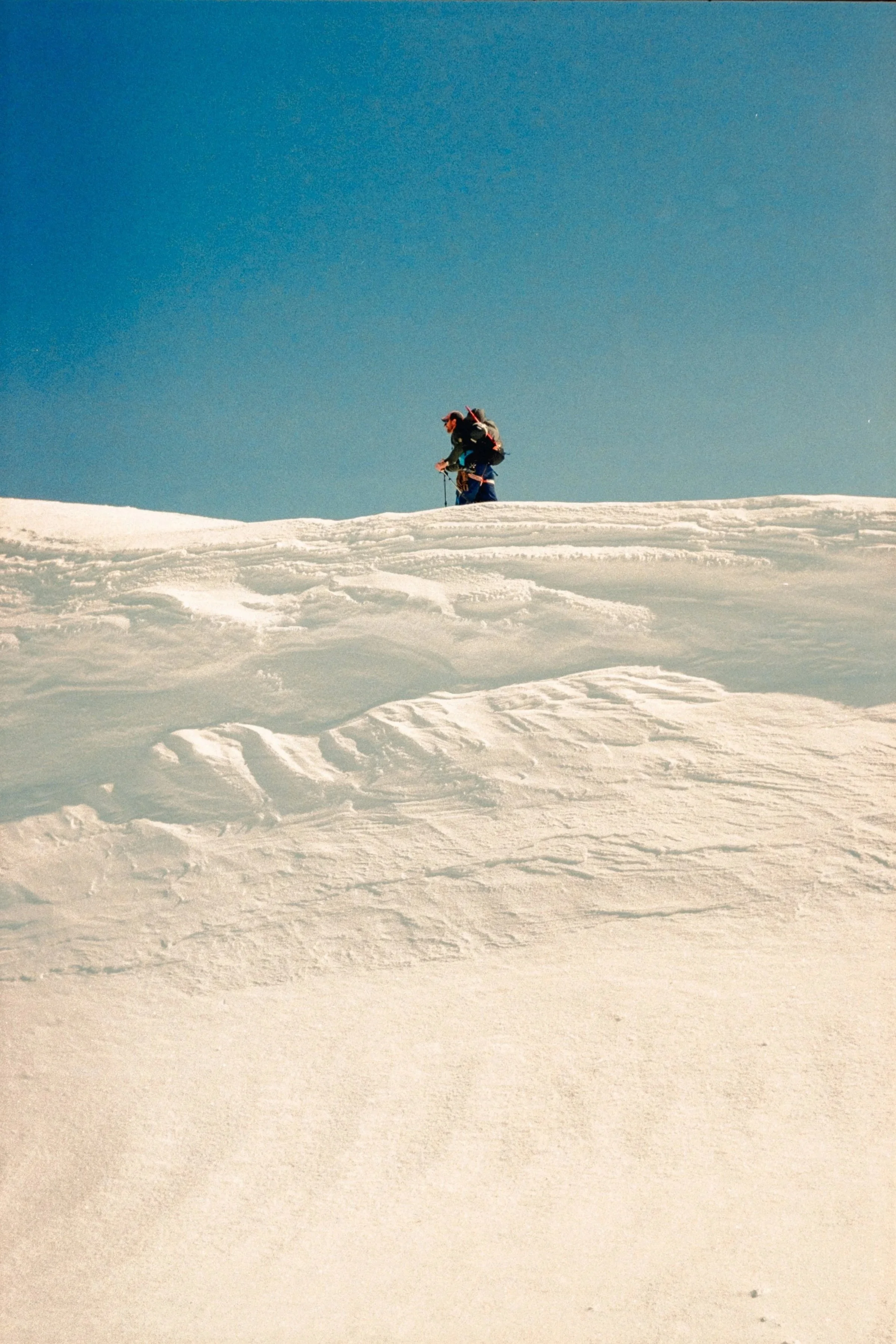 A hiker with a backpack and trekking poles walking on a snowy slope under a clear blue sky.