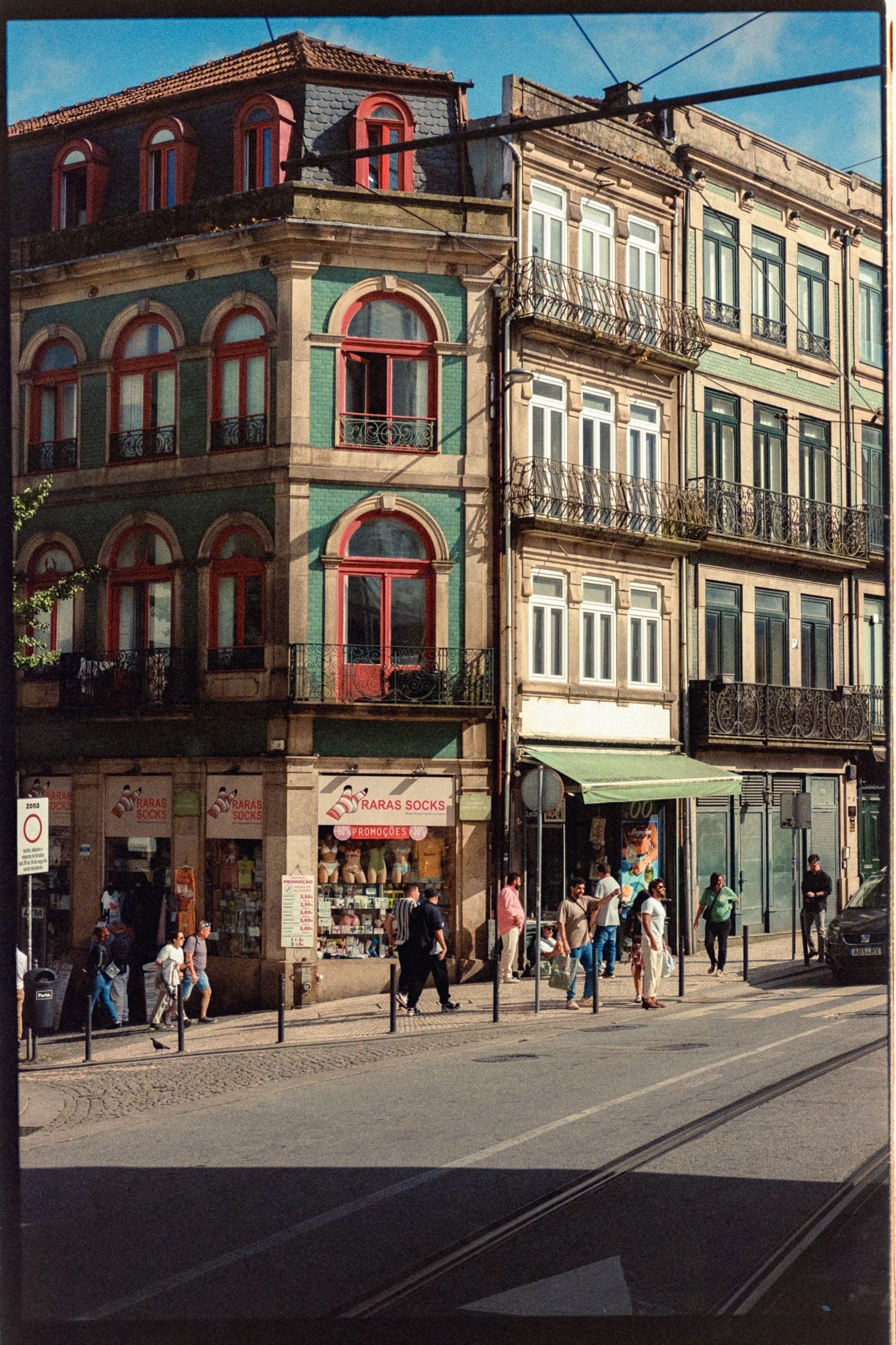 A street scene with pedestrians walking past shops on the ground floor of multi-story buildings with ornate balconies, large windows, and colorful facades, under a blue sky.