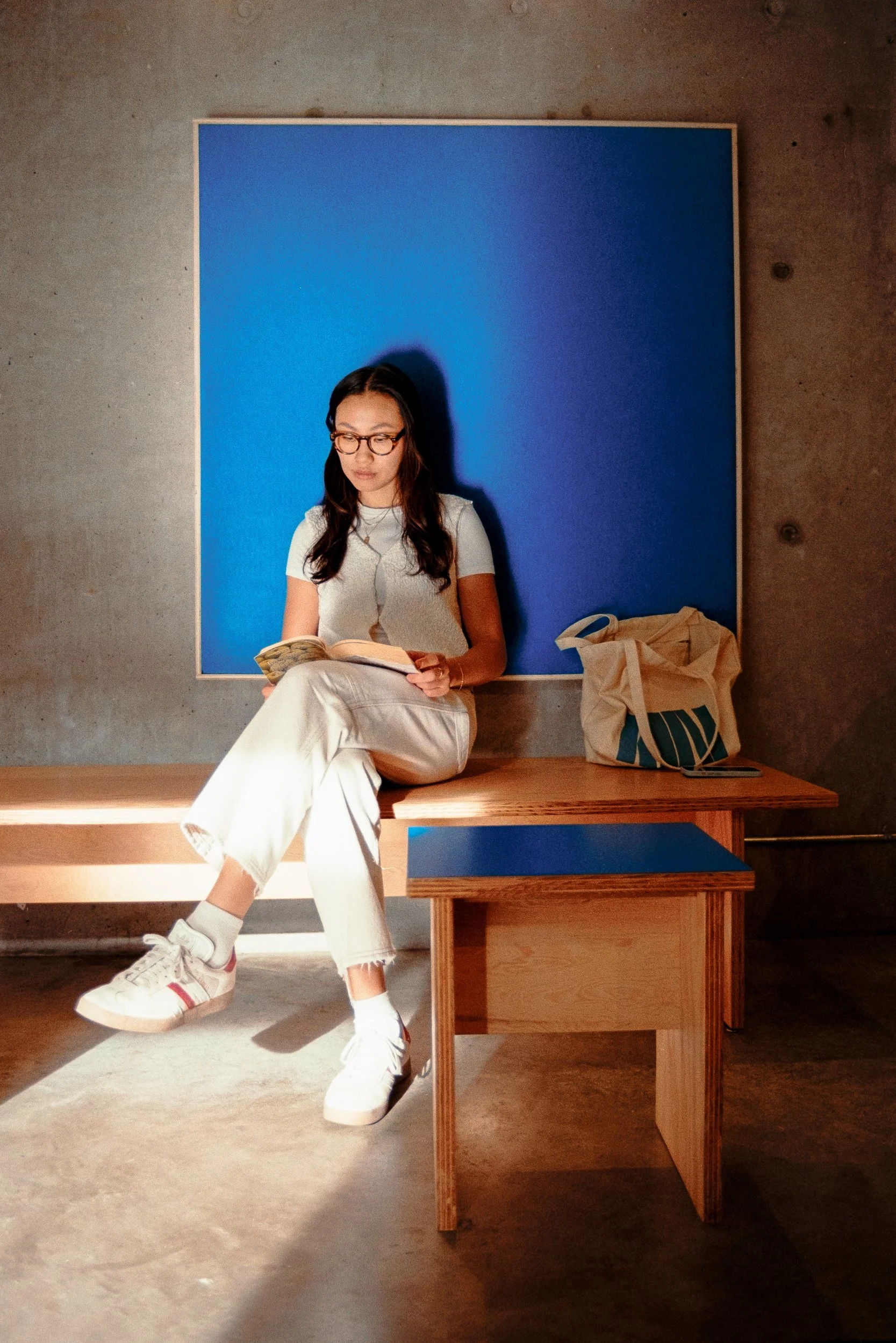 A young woman with glasses sitting on a wooden bench, reading a book, with a blue wall in the background and a beige tote bag on the bench beside her.
