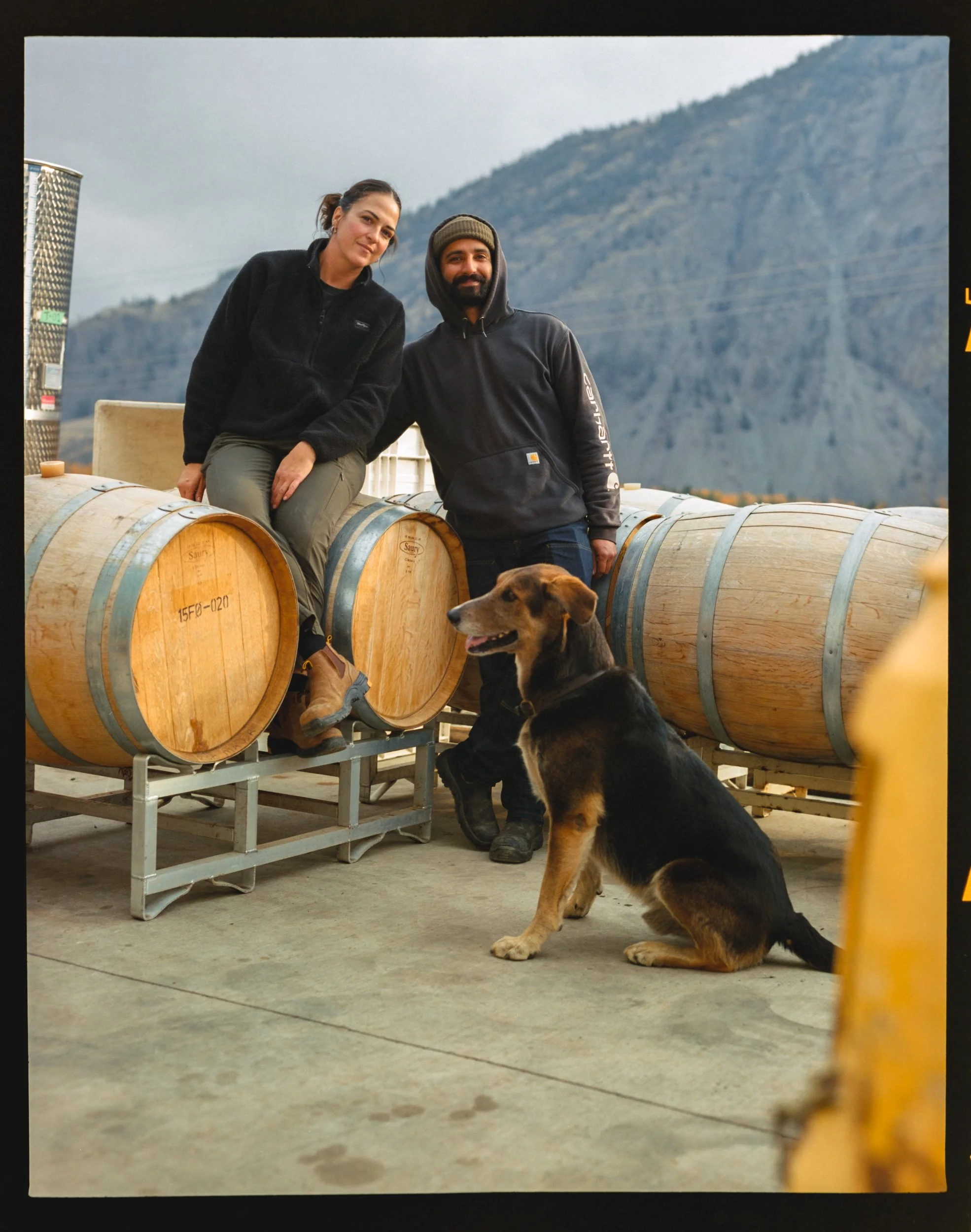 Two people, a woman and a man, are sitting and standing on large wooden wine barrels outdoors with a mountain in the background. A dog is sitting on the ground in front of them.