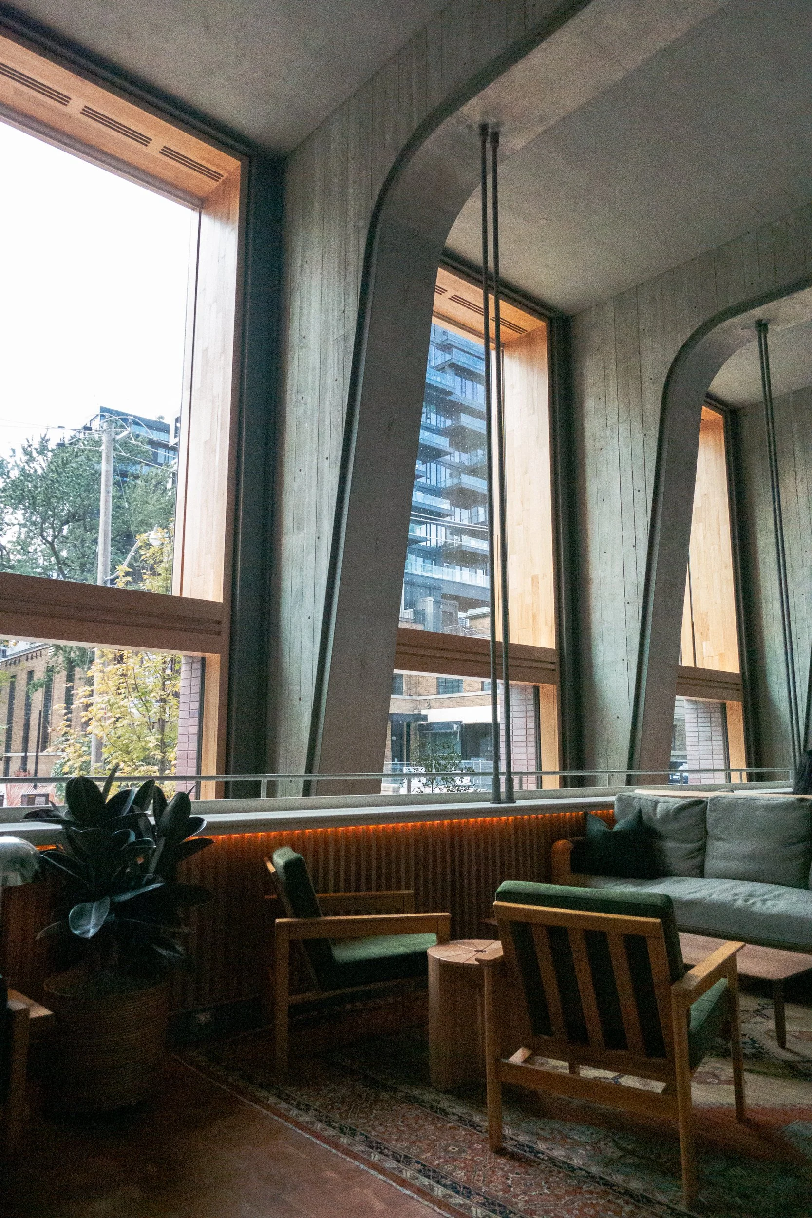 Interior of a modern room with large wooden framed windows, concrete walls, a potted plant, a vintage-style rug, and furniture including a sofa and chairs.