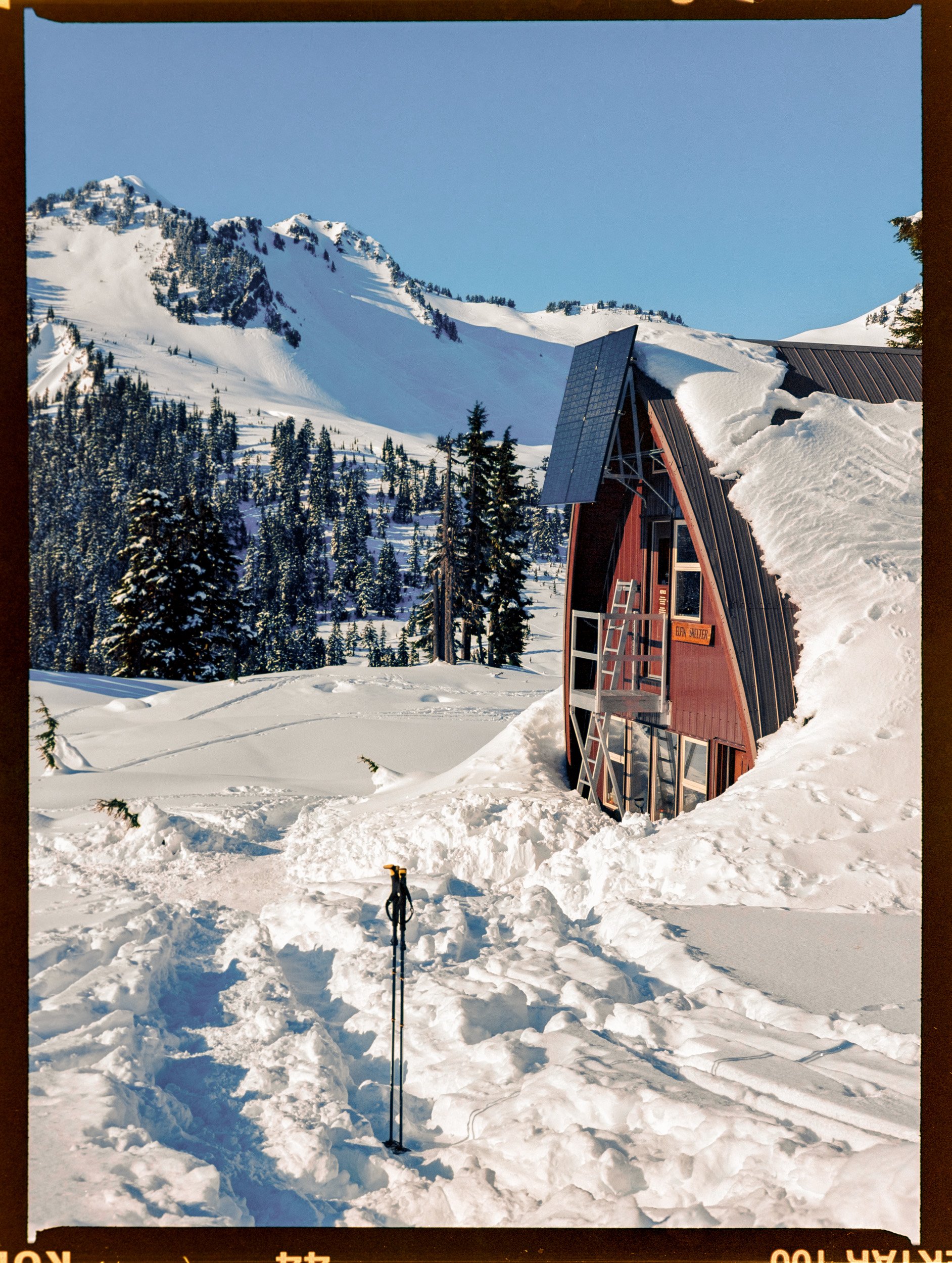 A-frame cabin with solar panel on snowy mountainside with forest and ski pole in foreground.