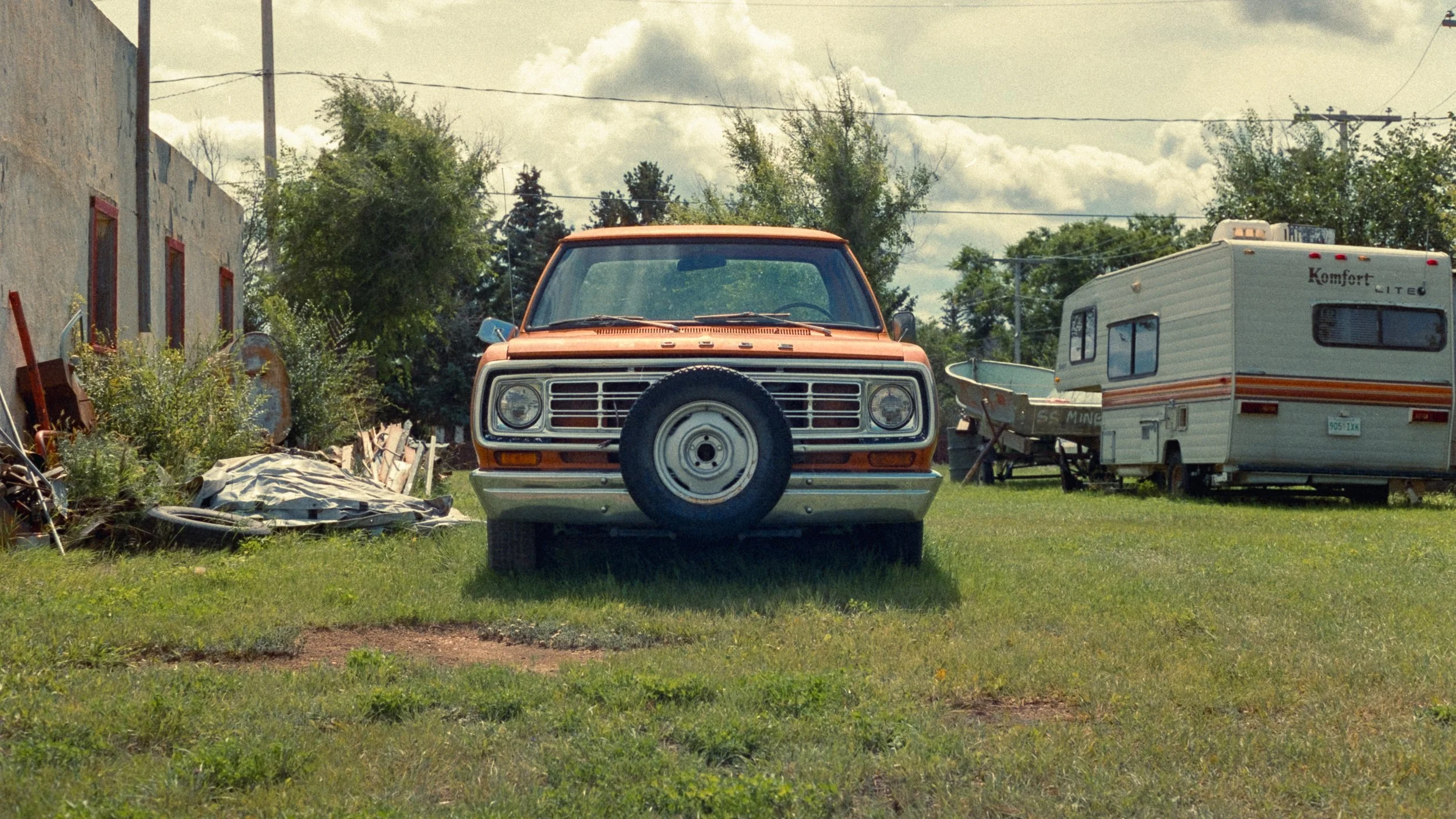 A vintage rust-colored Ford truck parked on a grassy lot with trees and a cloudy sky in the background. To the right, there is a white camper trailer with orange and grey stripes and a boat on a trailer nearby.