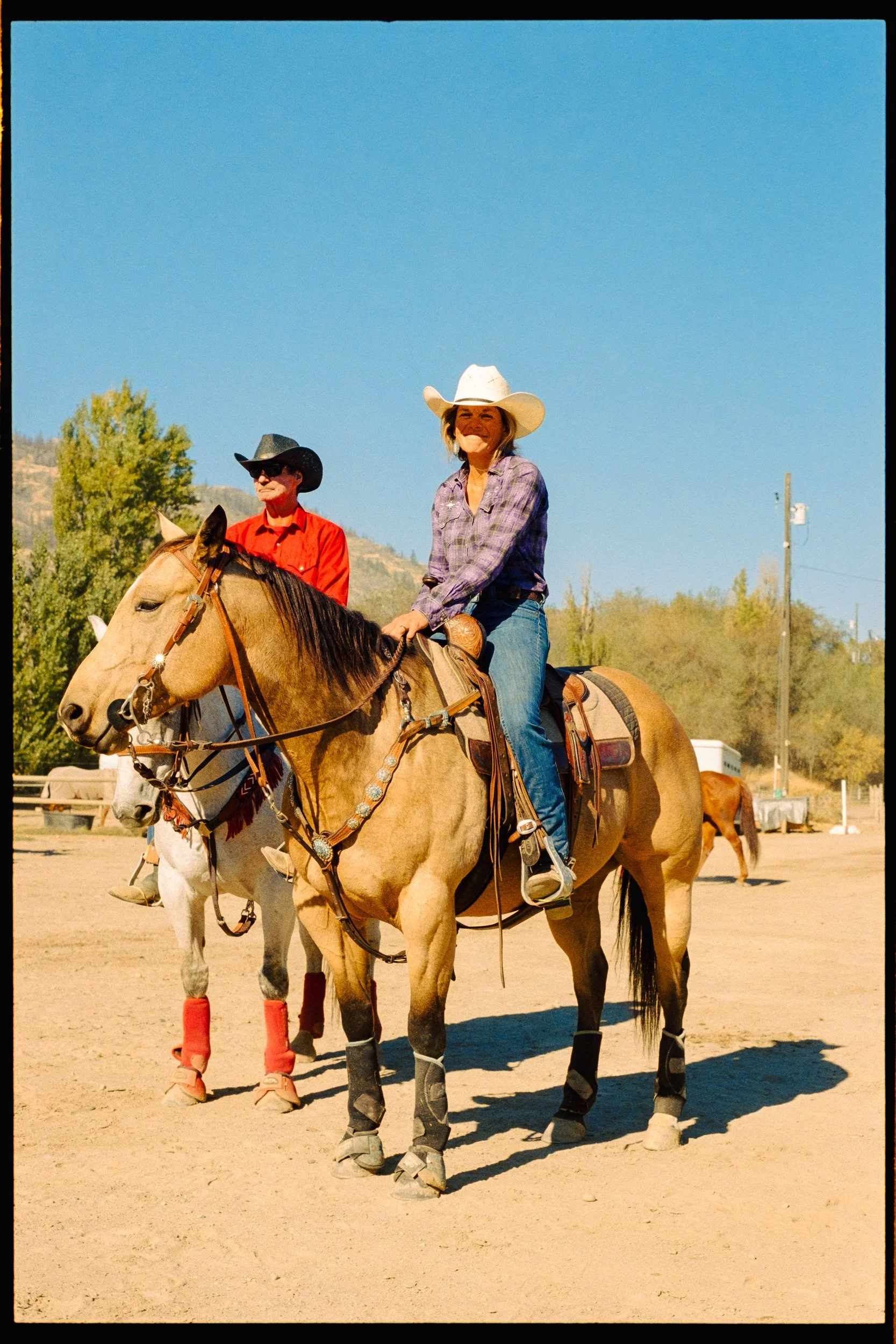 Two women riding horses outdoors under a clear blue sky, one wearing a white cowboy hat and plaid shirt, the other wearing a black cowboy hat and red shirt. Both horses are equipped with saddles and protective leg gear.