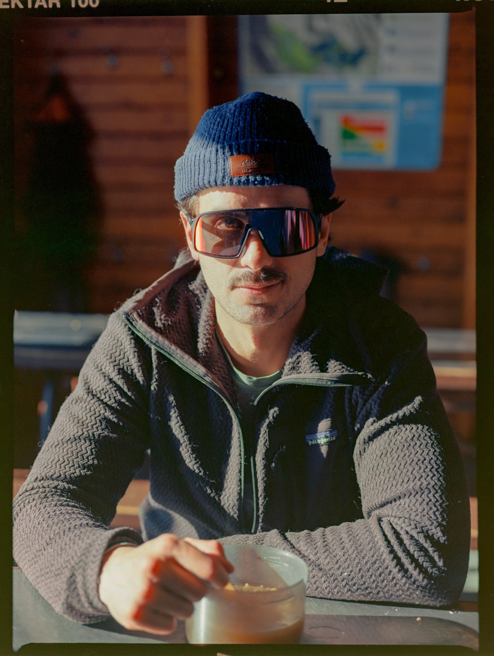 Person wearing sunglasses and a beanie sitting indoors, holding a bowl.
