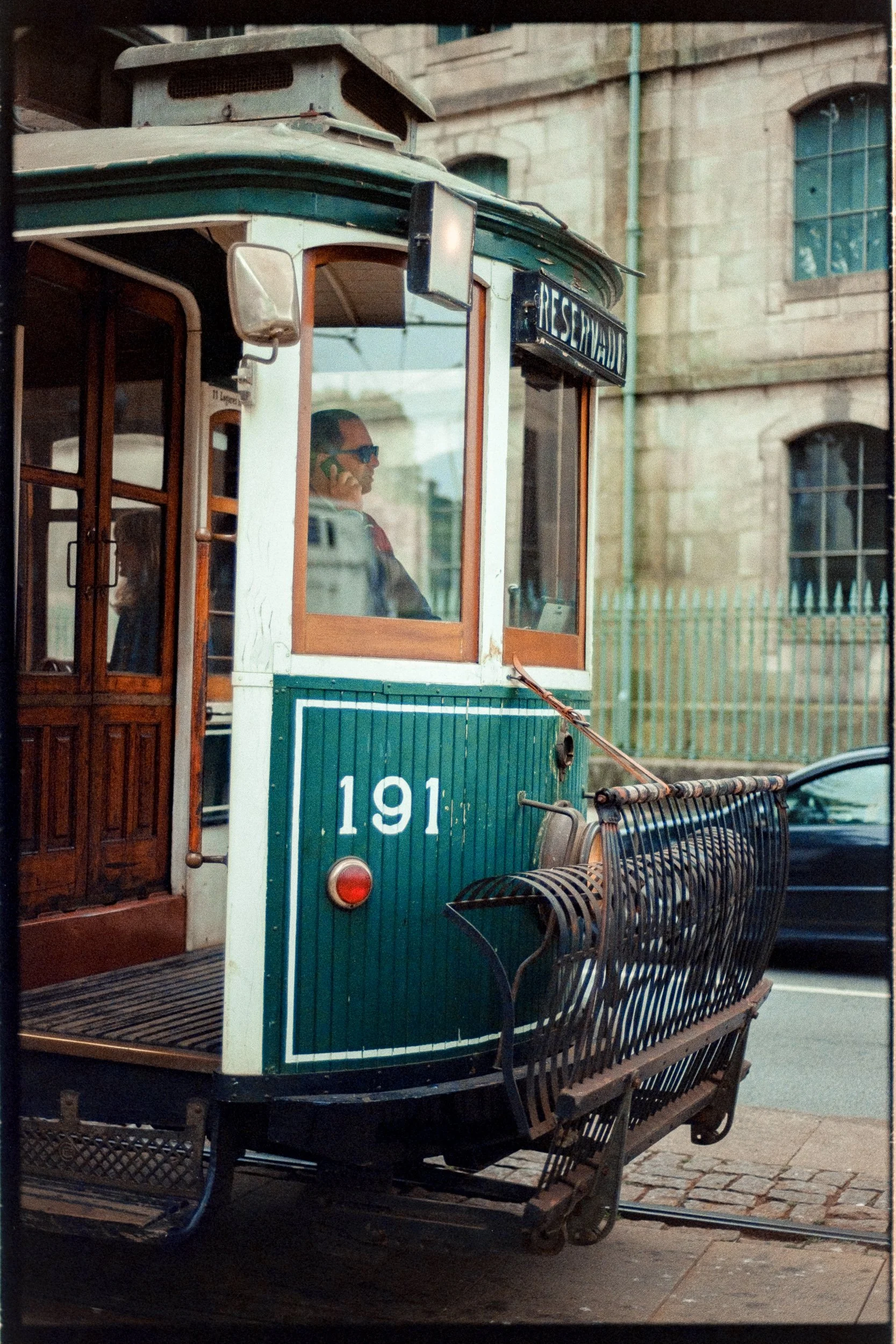 Vintage green and white tram with wooden accents, number 191, on a city street, with a person wearing sunglasses and talking on a cellphone inside, a metal bench at the tram's front, and historic stone buildings with large windows in the background.