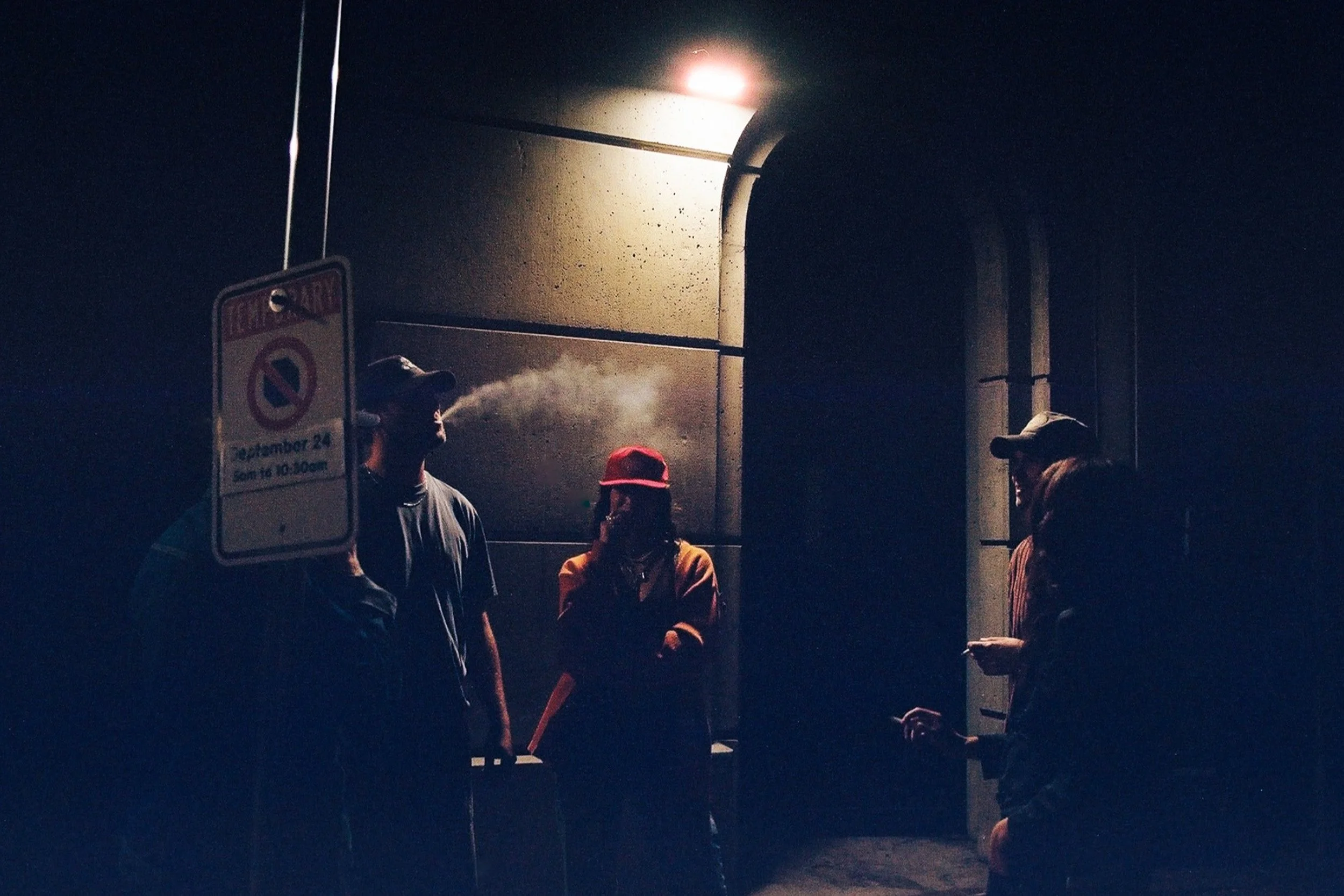 Four people standing under a streetlamp at night. One person is exhaling vapor or smoke, and another is on their phone. There's a no parking sign next to them.