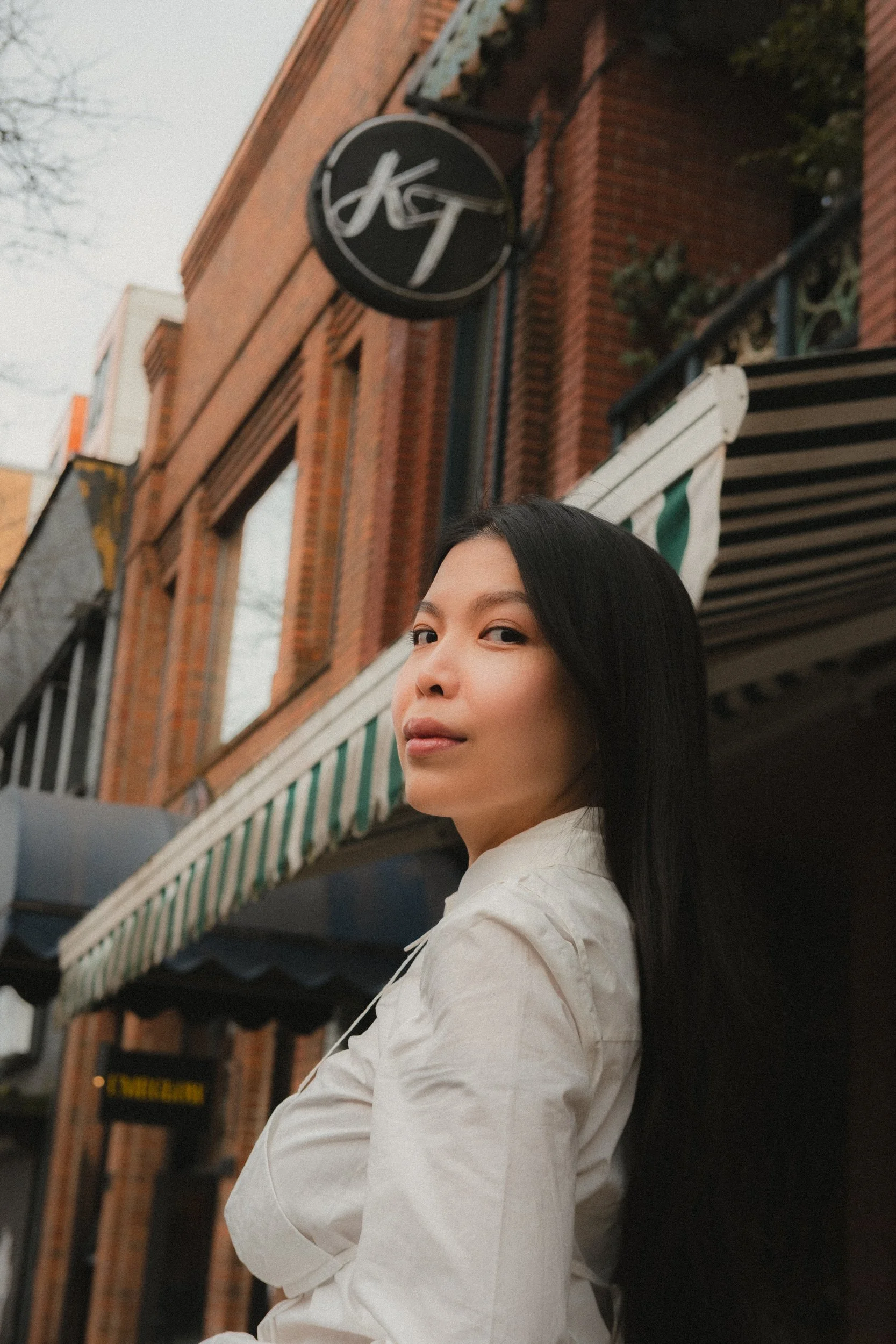 A woman with long black hair wearing a white shirt standing outside a brick building with green and white striped awning and a black round sign with white writing.