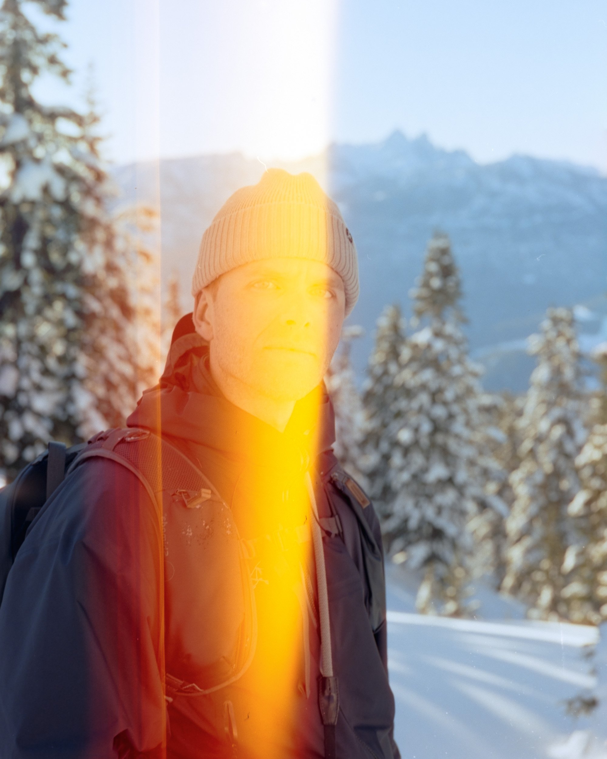 A person wearing a beanie and jacket standing in a snowy mountainous landscape with evergreen trees. There is a vertical light leak across the image.