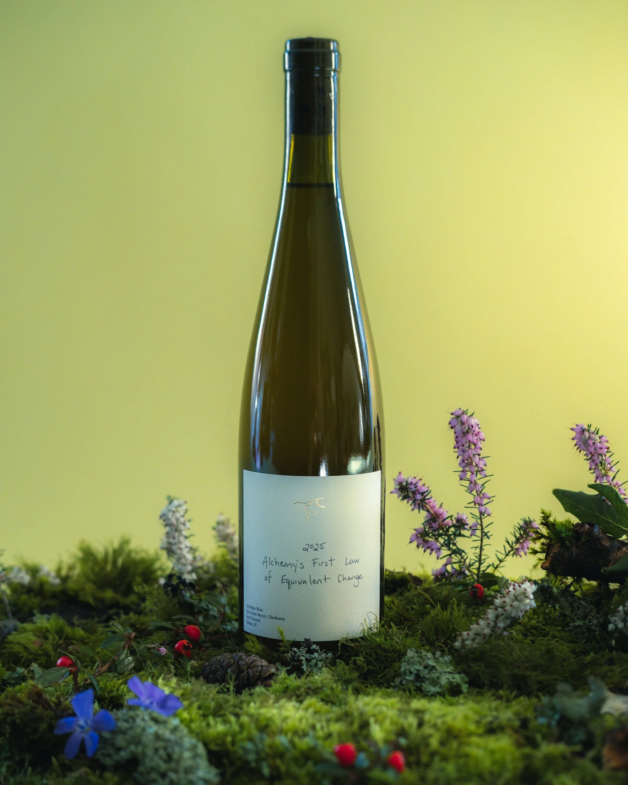 A wine bottle with a white label that reads '2025 Alchemy's First Law of Equivalent Exchange,' placed on a bed of moss, flowers, and pinecones, with a yellow background.
