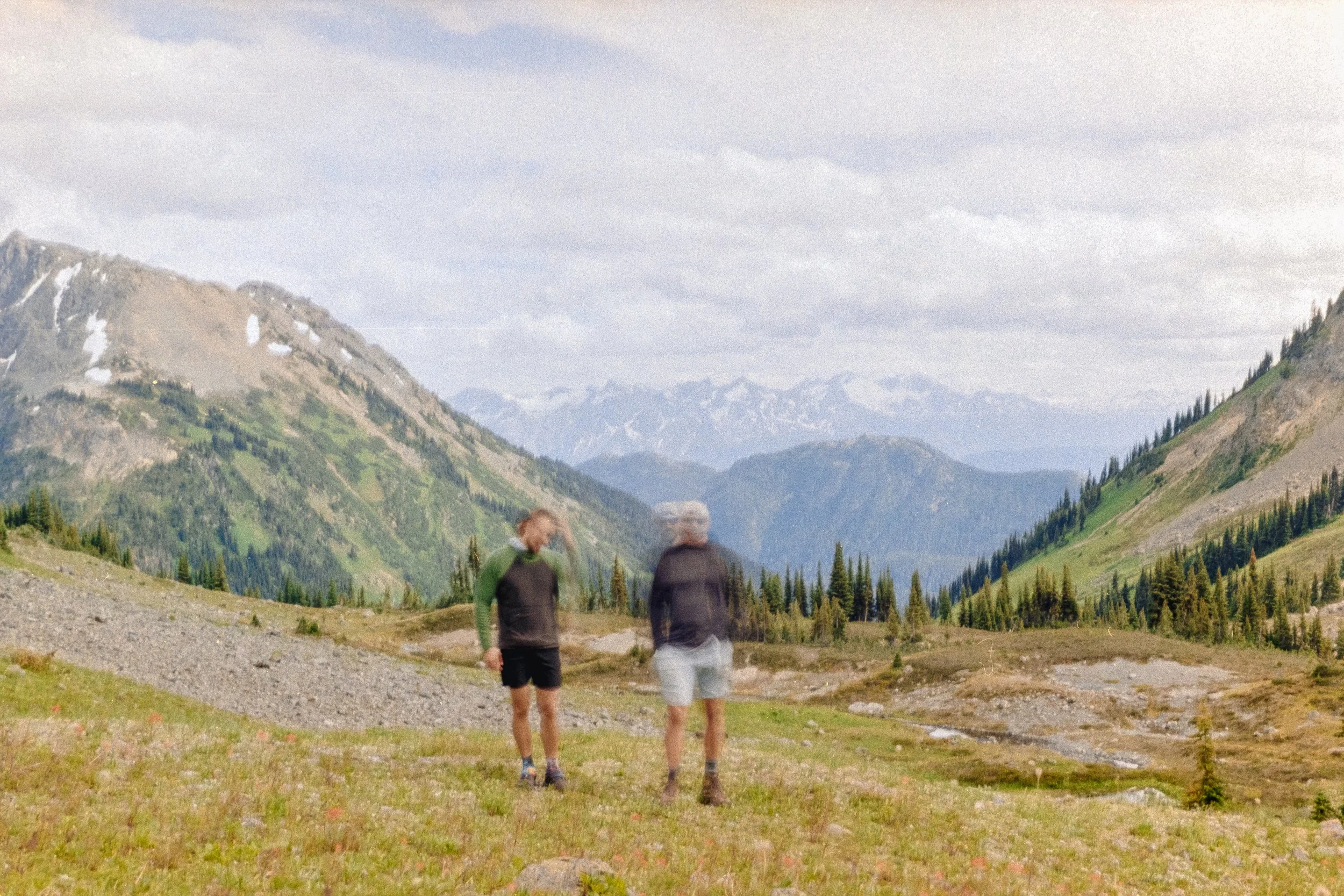 Two people hiking in a mountainous landscape with green hills, trees, and snow-capped peaks in the background, under a cloudy sky.