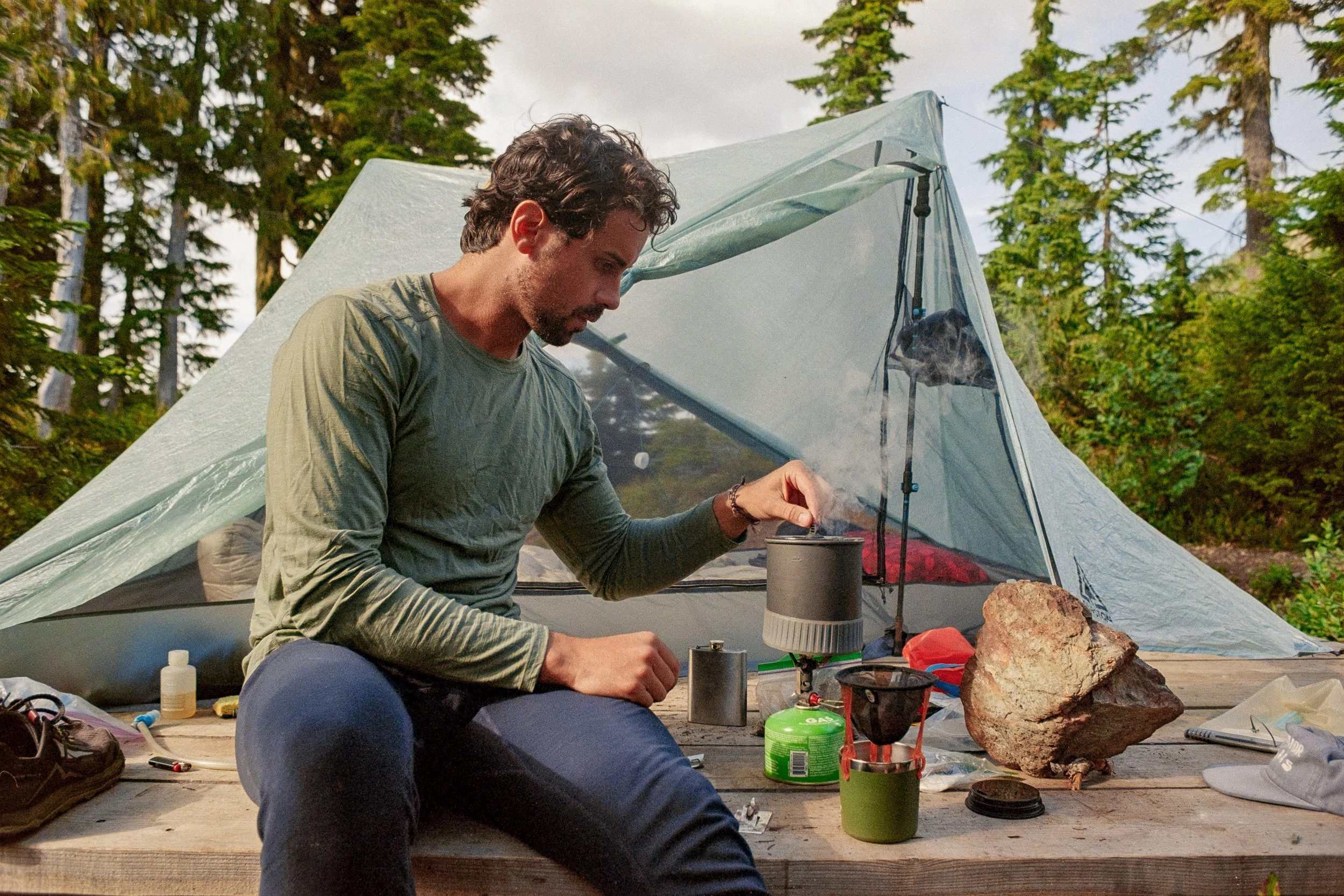 A man preparing a camping stove outdoors near a small tent in a forest.