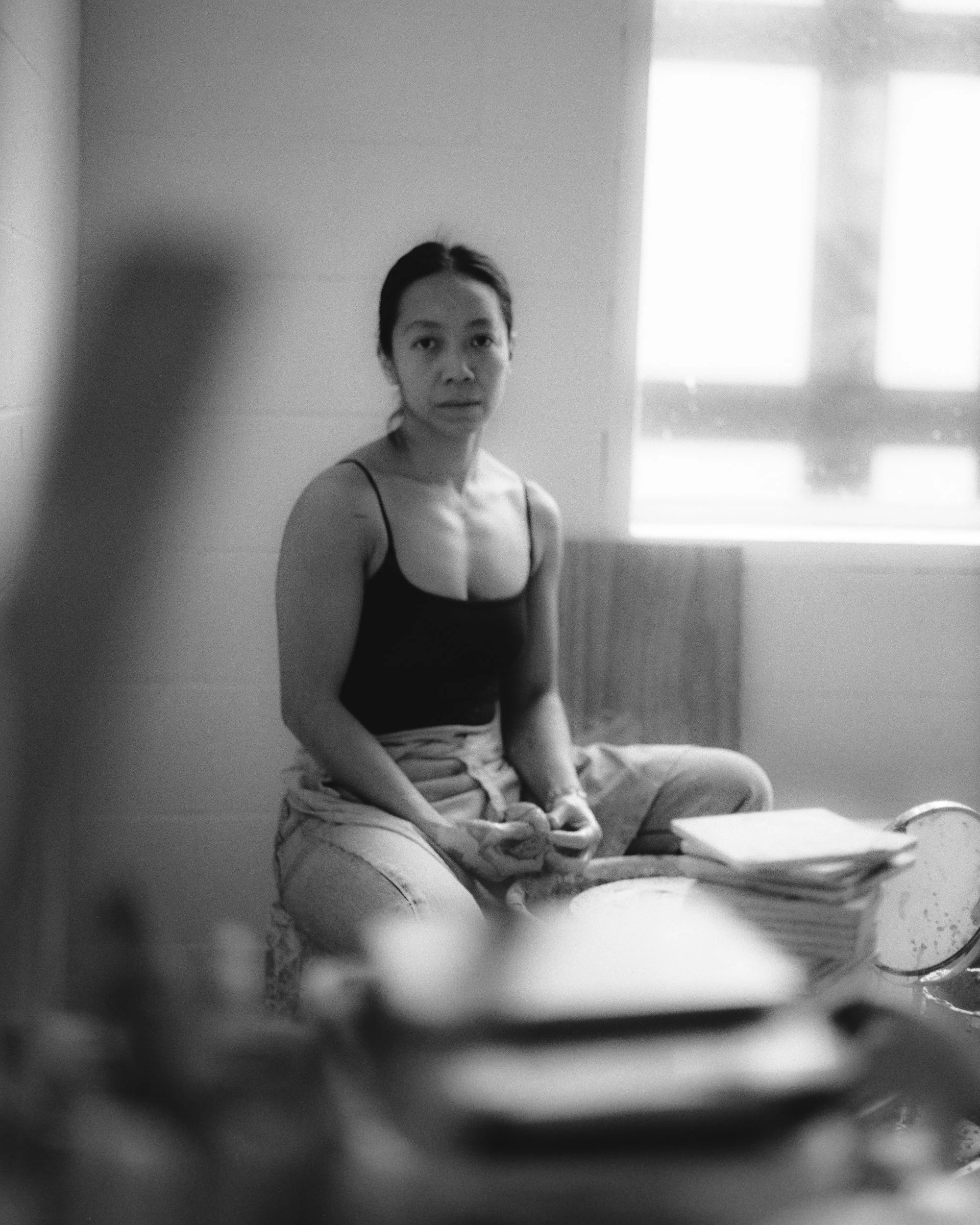 A woman sits on a chair in a room with a window in the background, looking directly at the camera with a neutral expression.