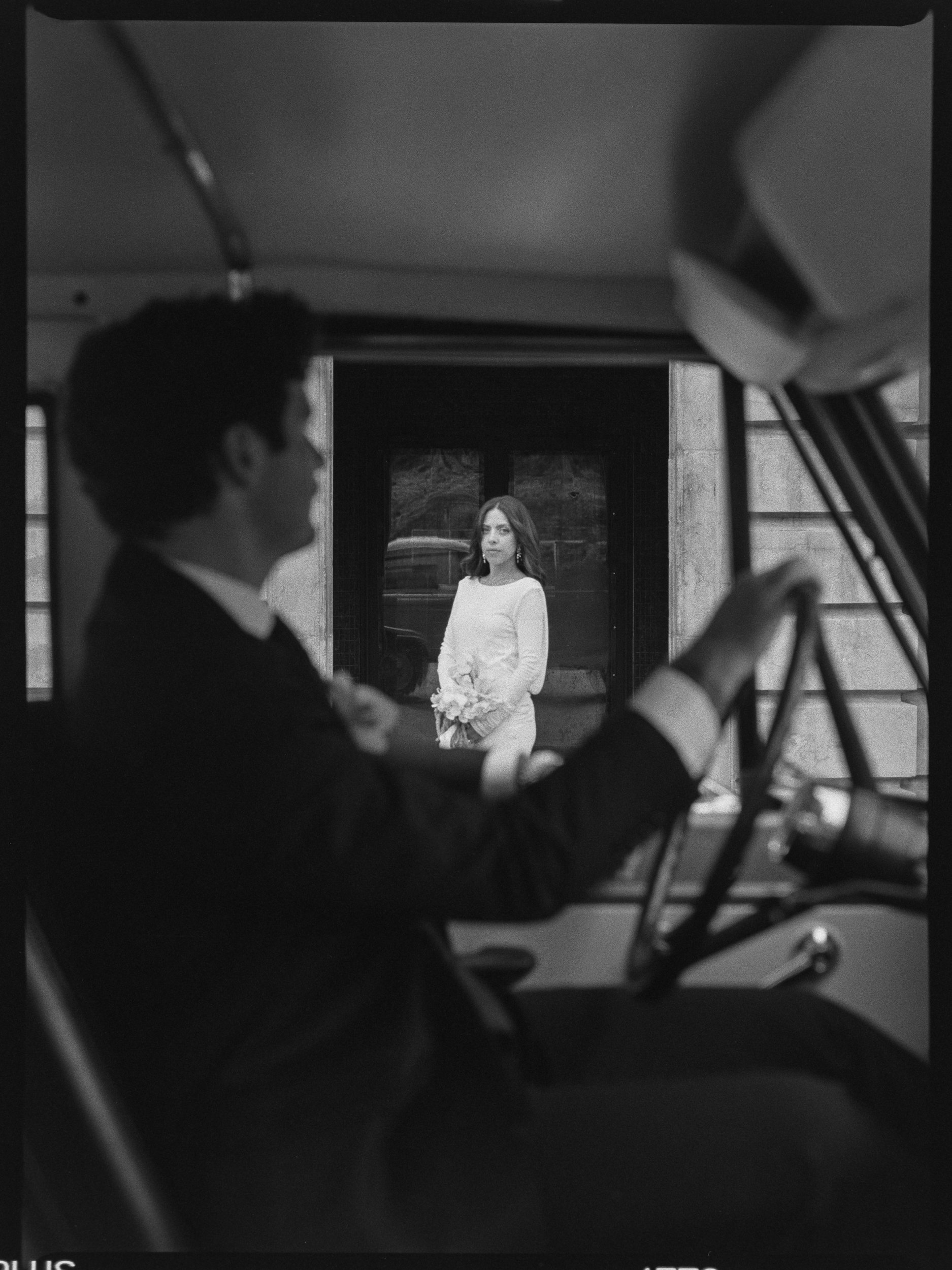 A man in a suit driving a bus, with a woman in a white dress holding a bouquet standing outside the window and looking inside.