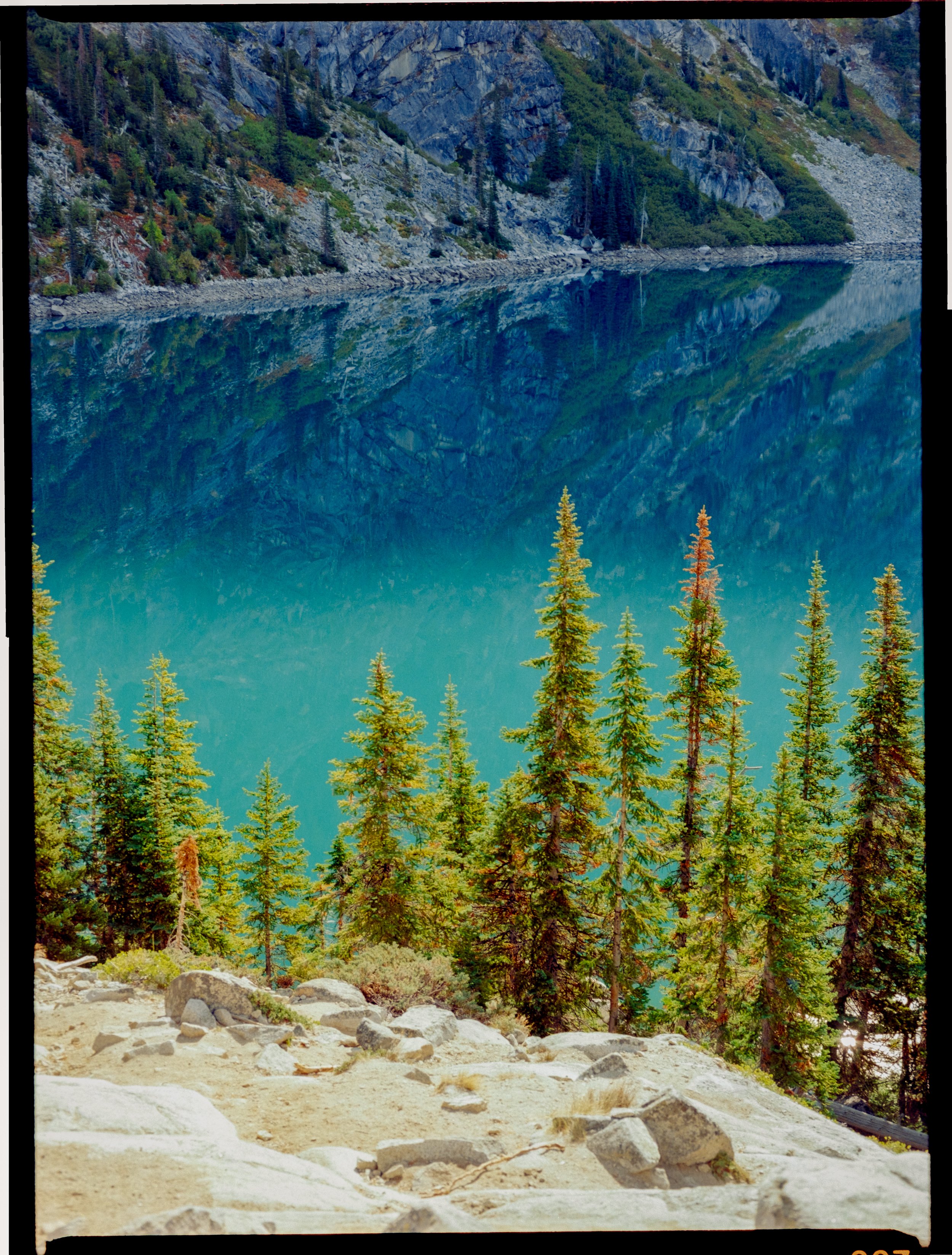 A scenic view of tall evergreen trees by a clear, reflective blue lake with rocky mountains in the background.