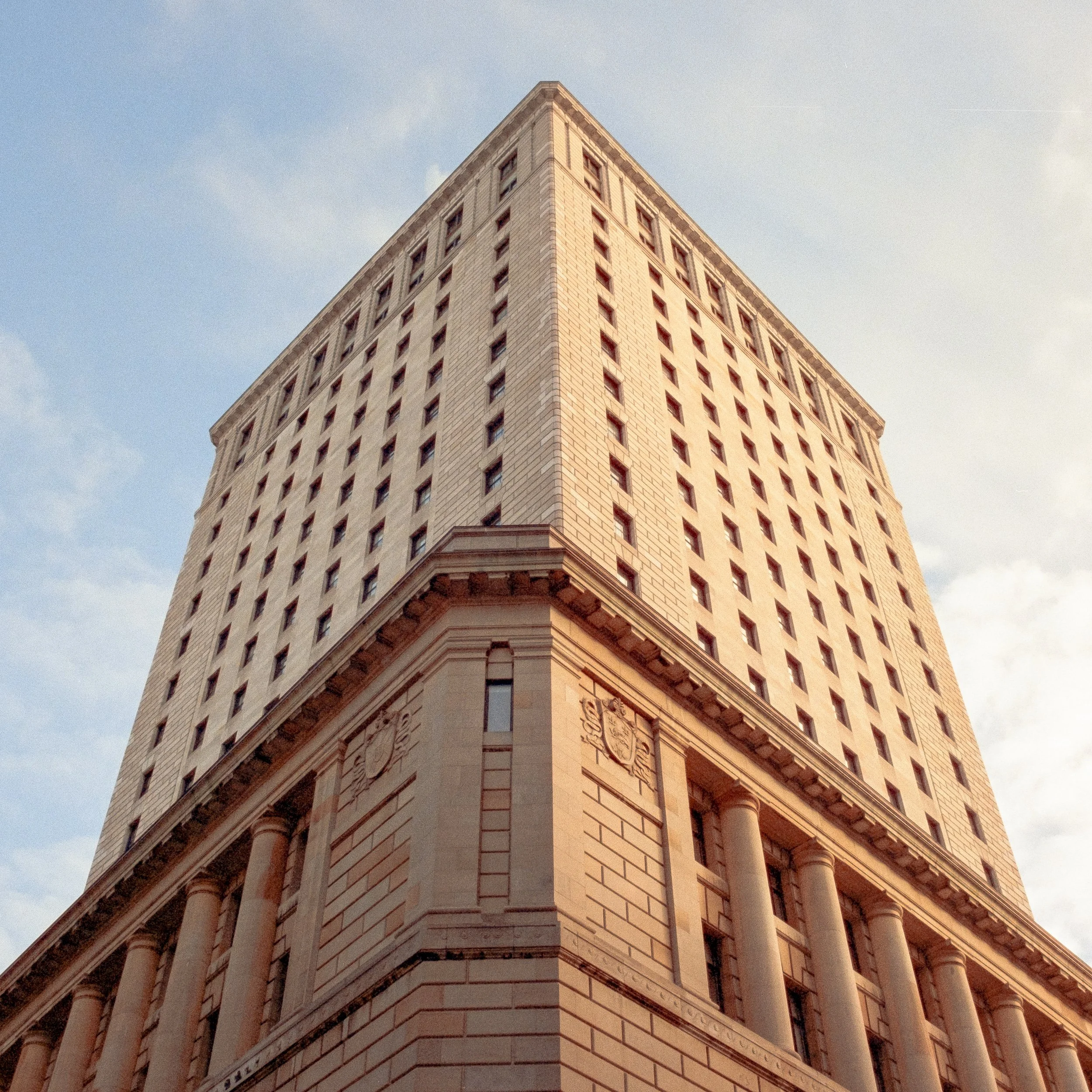 A tall, historic building with a stone facade, columns, and decorative emblems, seen from a low angle against a blue sky with some clouds.