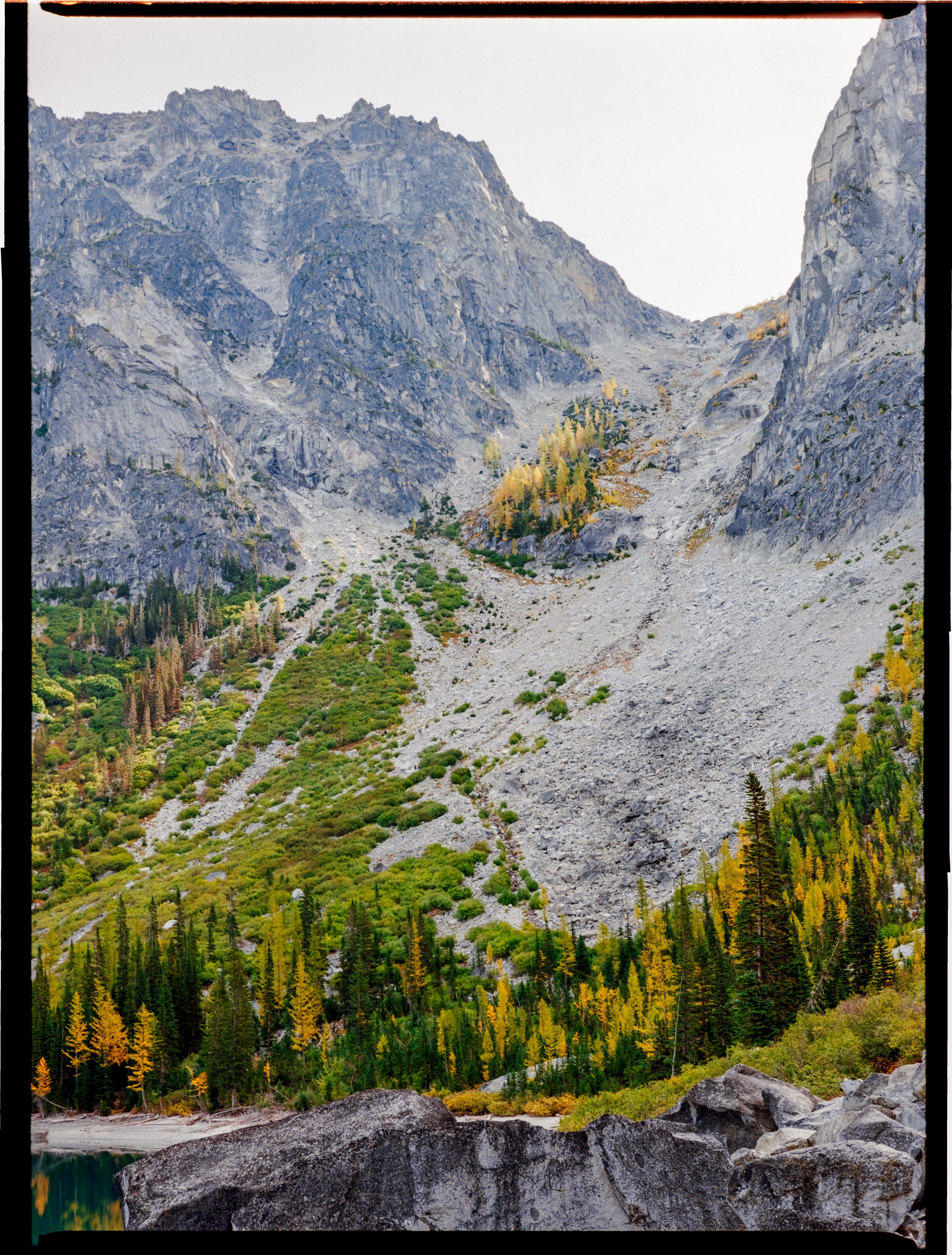 Rocky mountain landscape with green and yellow foliage, conifer trees, and a clear sky.