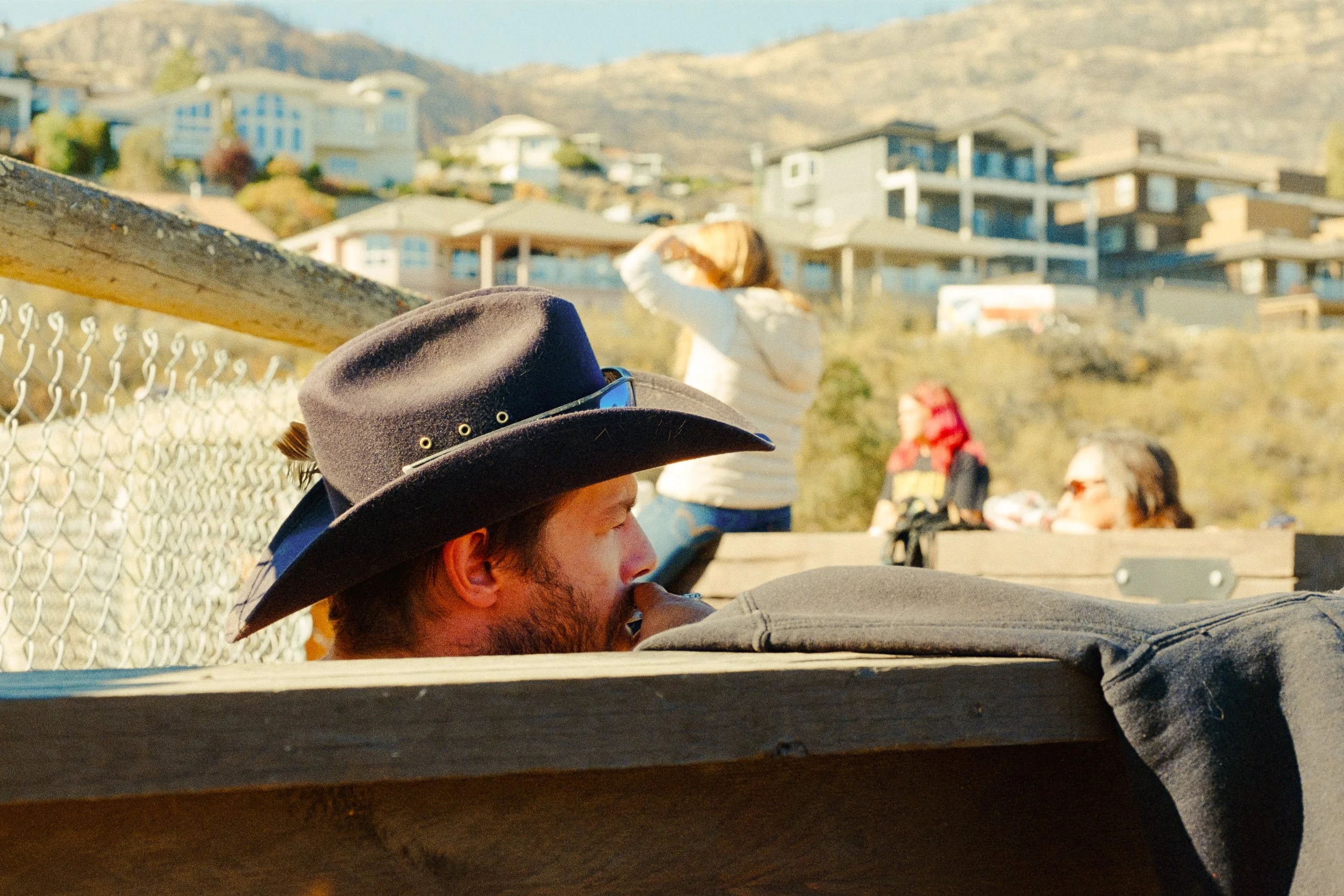 Side profile of a man with a beard wearing a gray hat, sitting outdoors in front of a chain-link fence, with hillside houses in the background under clear sky.