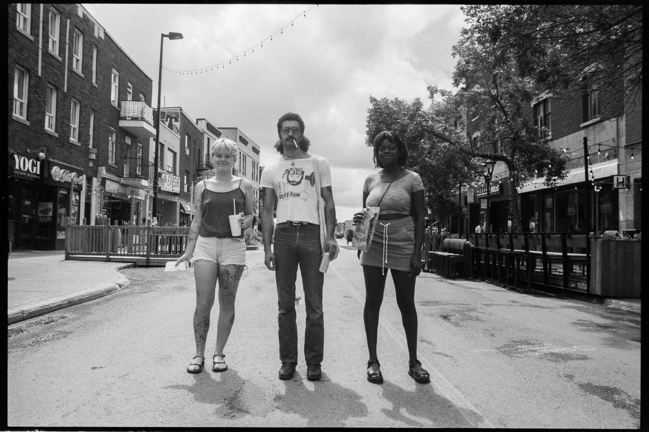 Three people stand on the middle of a city street in black and white. They are facing the camera, with buildings and trees in the background. The person on the left is a woman with short blonde hair, wearing a tank top and shorts, holding a drink wit