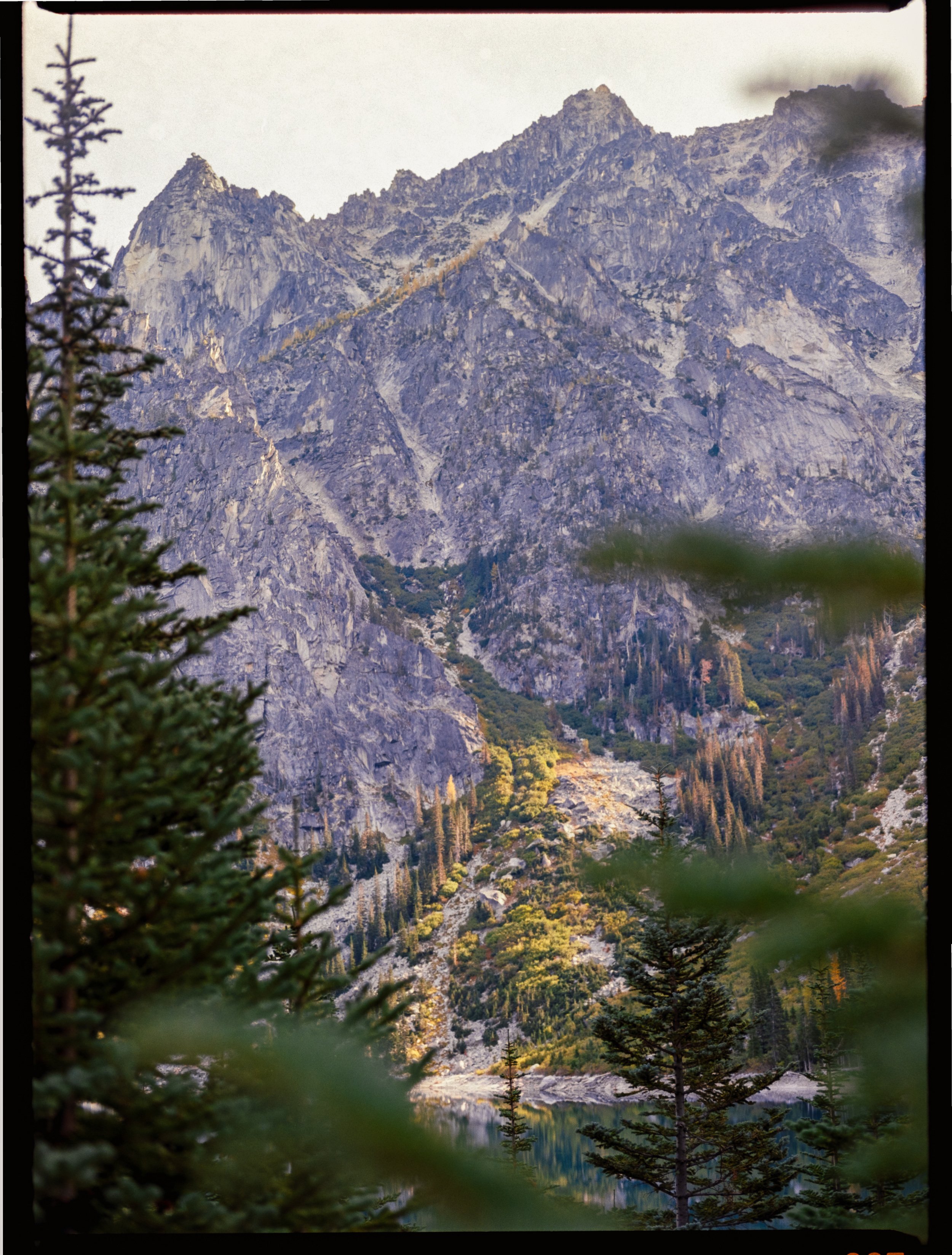 Mountain landscape with rocky peaks, scattered evergreen trees, and a lake nestled below. The scene features lush green vegetation and a clear sky.