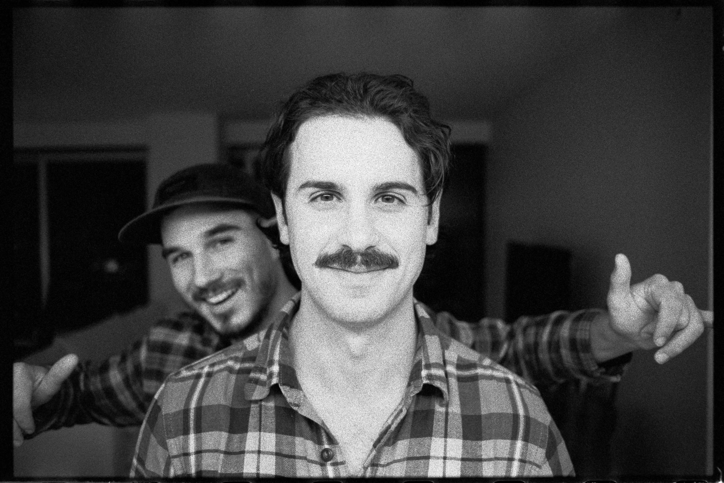 Two young men in casual checkered shirts, smiling and making playful gestures, indoors with a dark background.