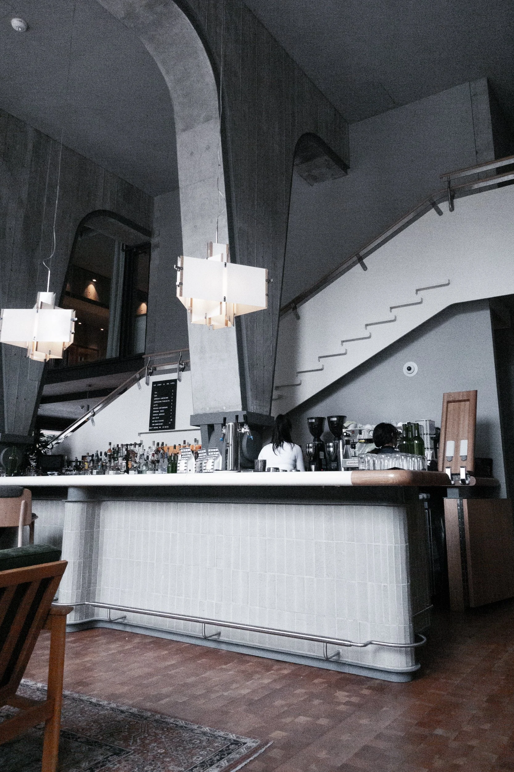 Modern bar with white countertop and hanging pendant lights, two staff members working behind the bar, shelves with bottles, staircase in the background, and contemporary architectural design.