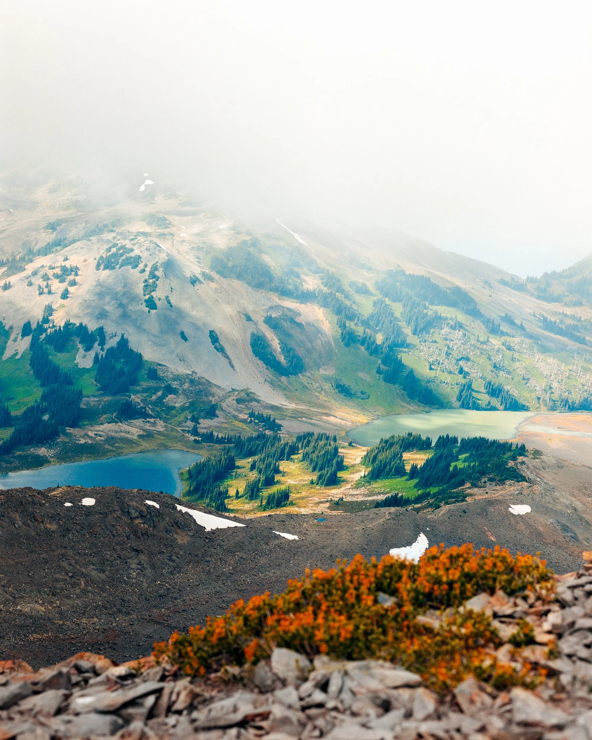 Aerial view of a mountain landscape with lakes, forests, and rocky terrain, partially obscured by low clouds or fog.
