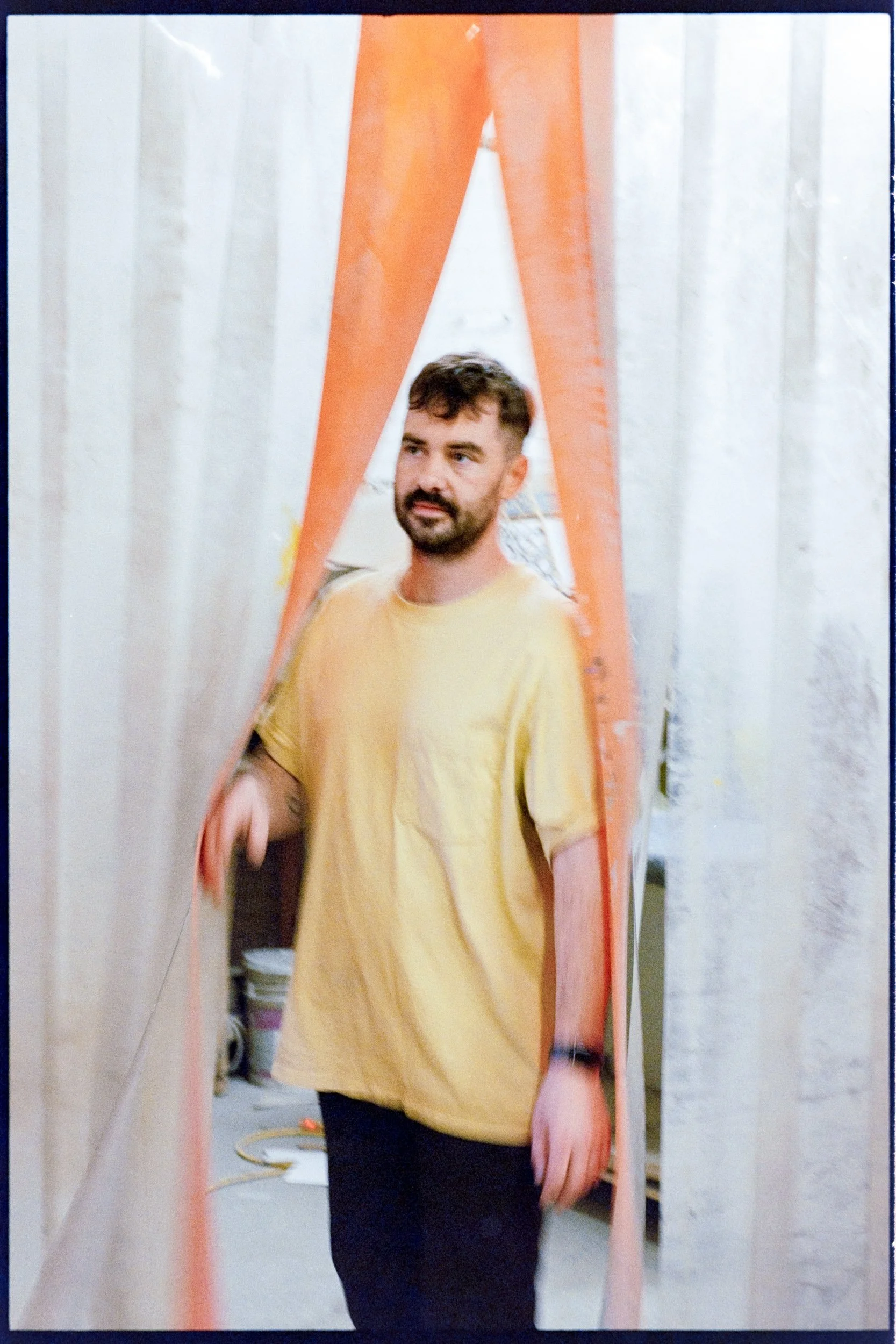 A man with a beard and short hair, wearing a yellow T-shirt and black pants, standing next to an orange fabric or curtain, in an indoor setting with light-colored walls and some clutter in the background.