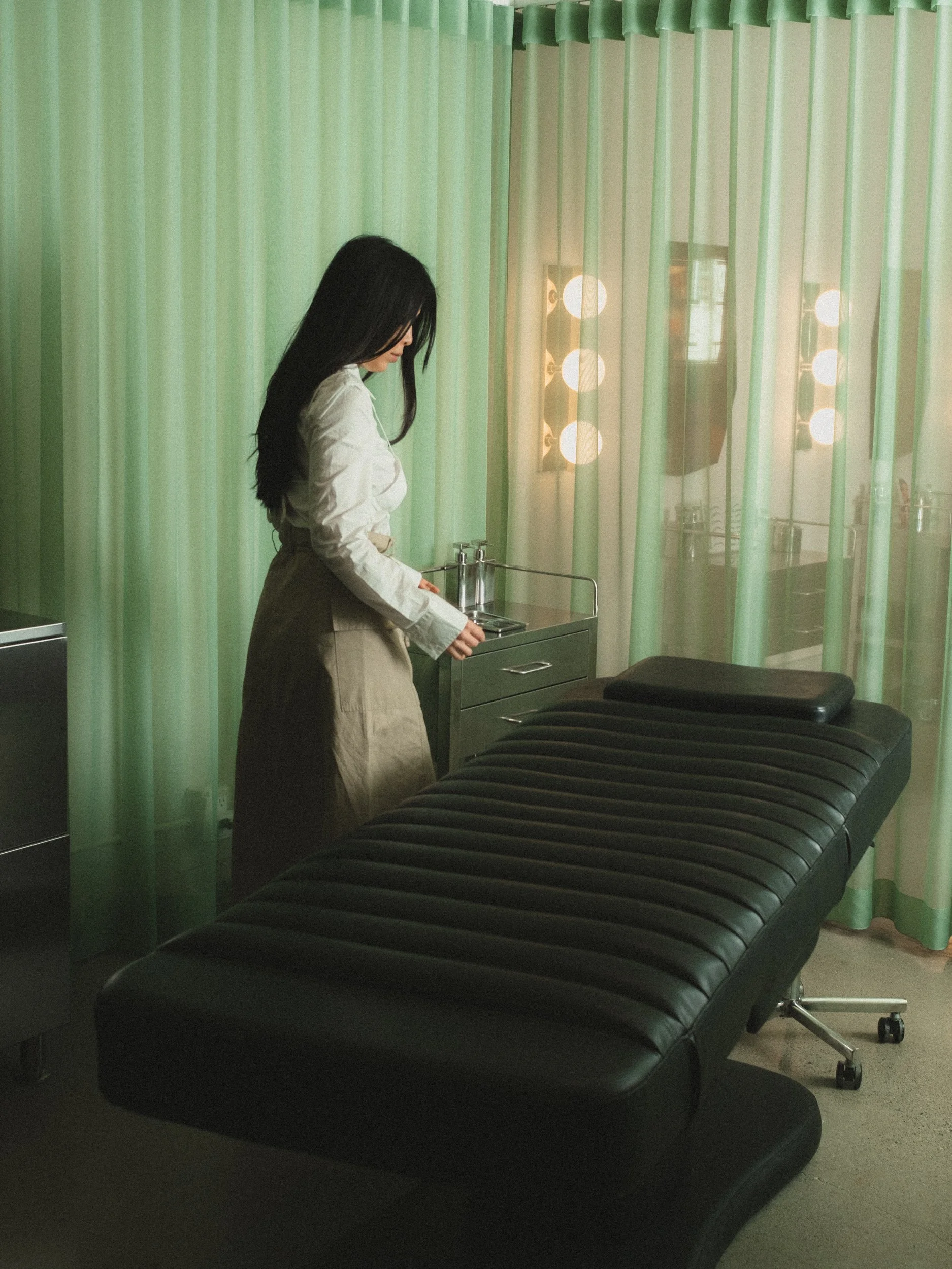 A woman standing beside a black massage or examination table in a room with green curtains and a mirror with light bulbs around it.